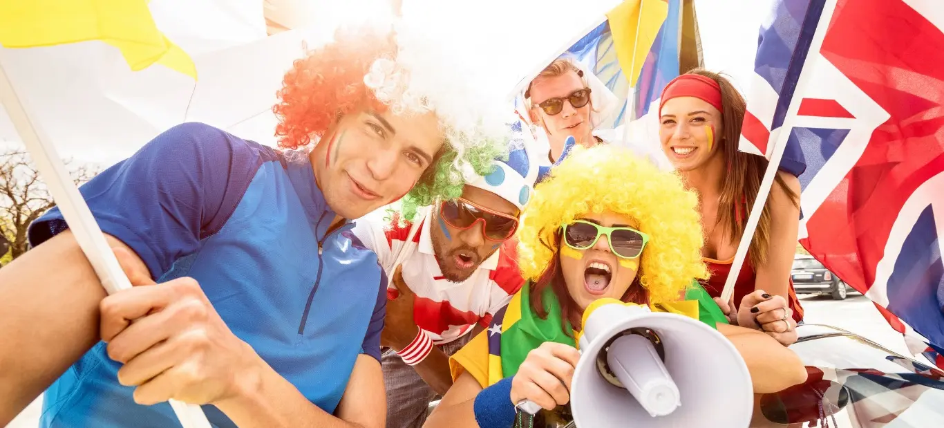 Five people wearing colorful wigs and face paint holding flags and a megaphone, cheering outdoors in bright sunlight.