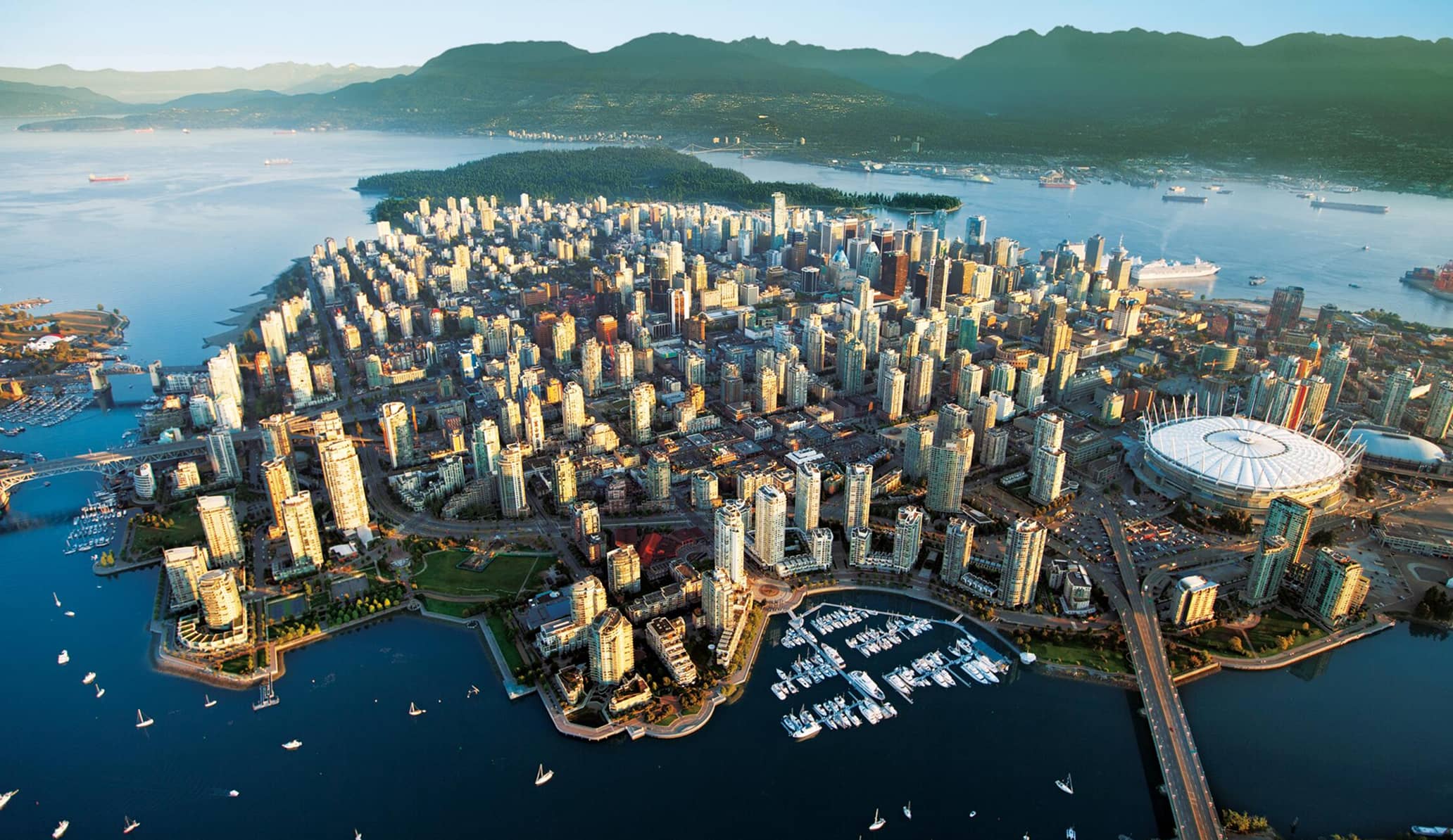 Aerial view of a coastal city with numerous high-rise buildings, a marina with boats, a stadium, and surrounding water with mountains in the background.