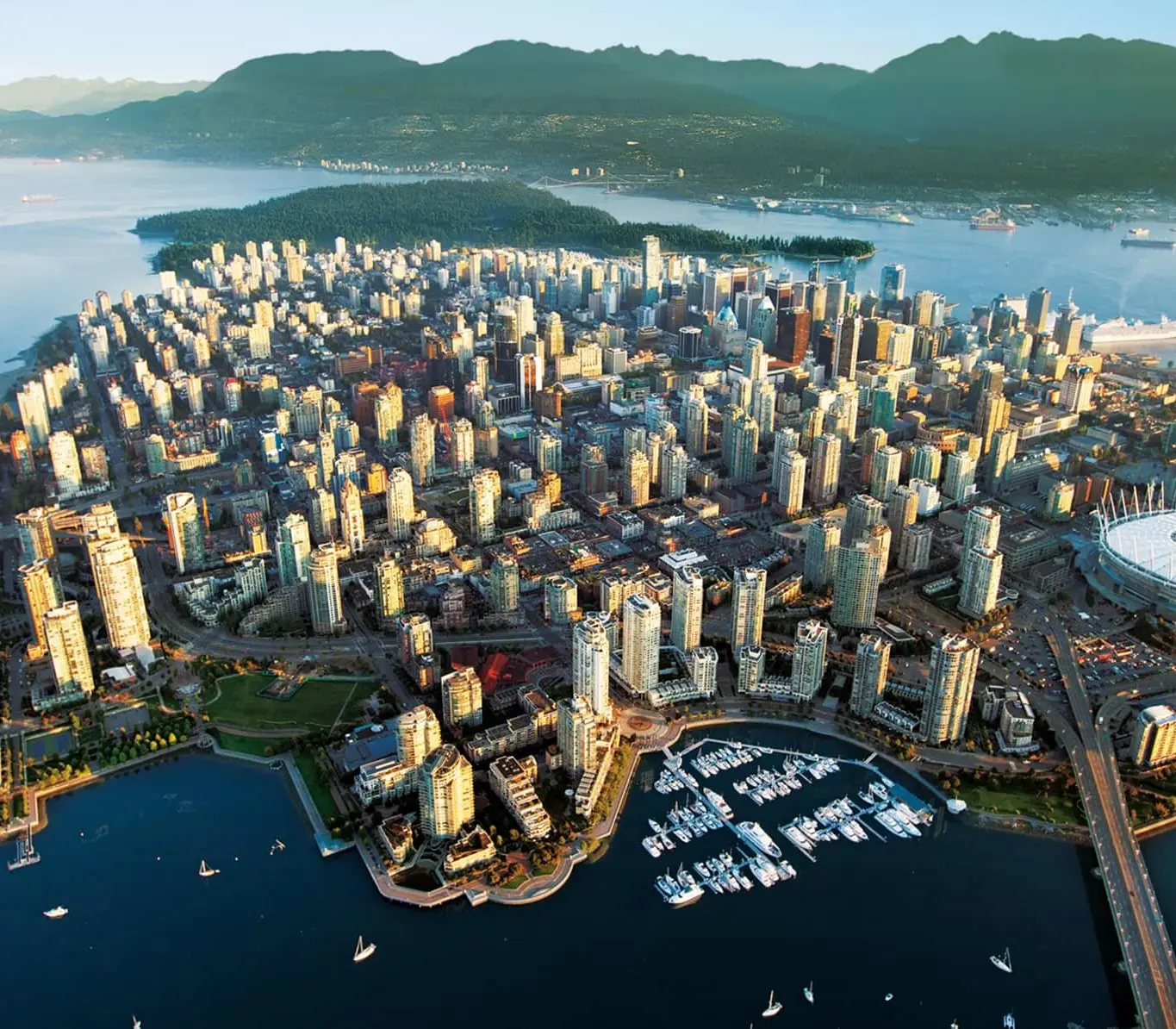 Aerial view of a coastal city with numerous high-rise buildings, a marina with boats, a stadium, and surrounding water with mountains in the background.