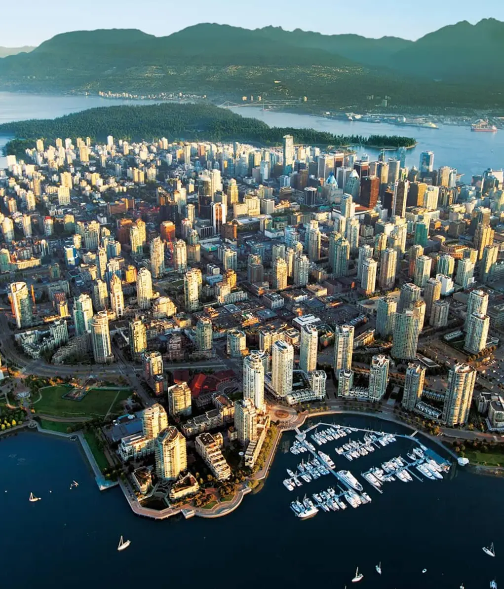 Aerial view of a coastal city with numerous high-rise buildings, a marina with boats, a stadium, and surrounding water with mountains in the background.