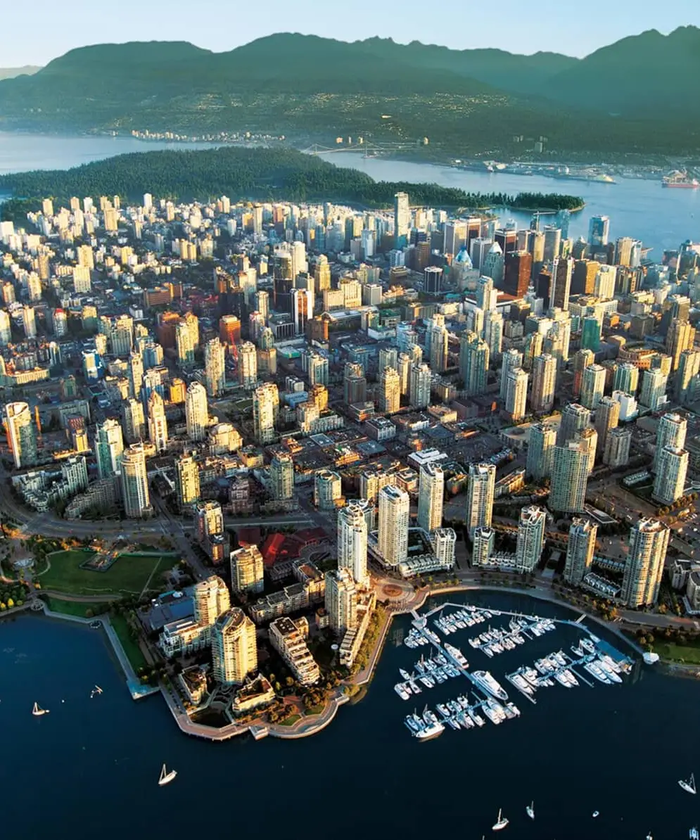 Aerial view of a coastal city with numerous high-rise buildings, a marina with boats, a large stadium, and surrounding water and mountains.