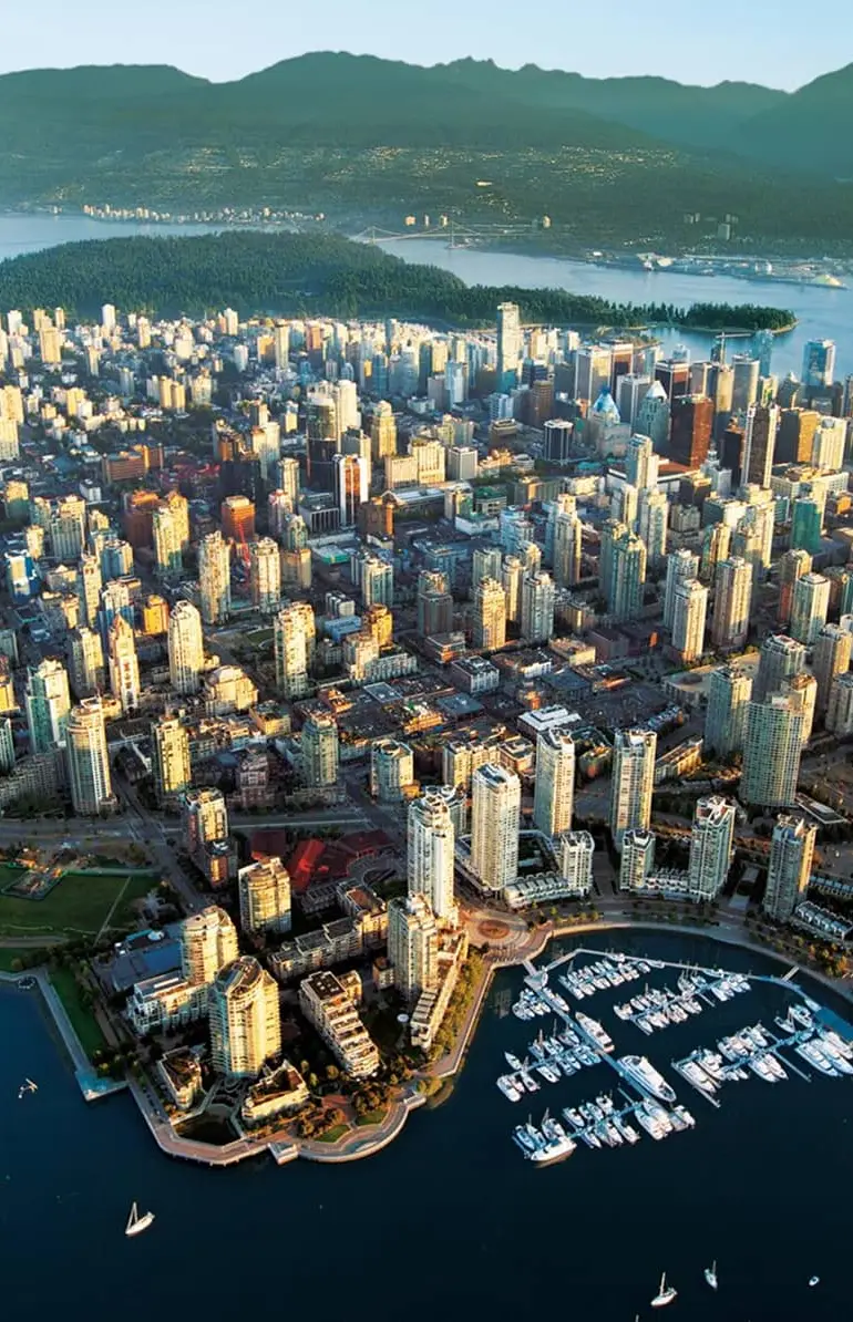 Aerial view of a coastal city with numerous high-rise buildings, a marina with boats, a stadium, and surrounding water with mountains in the background.