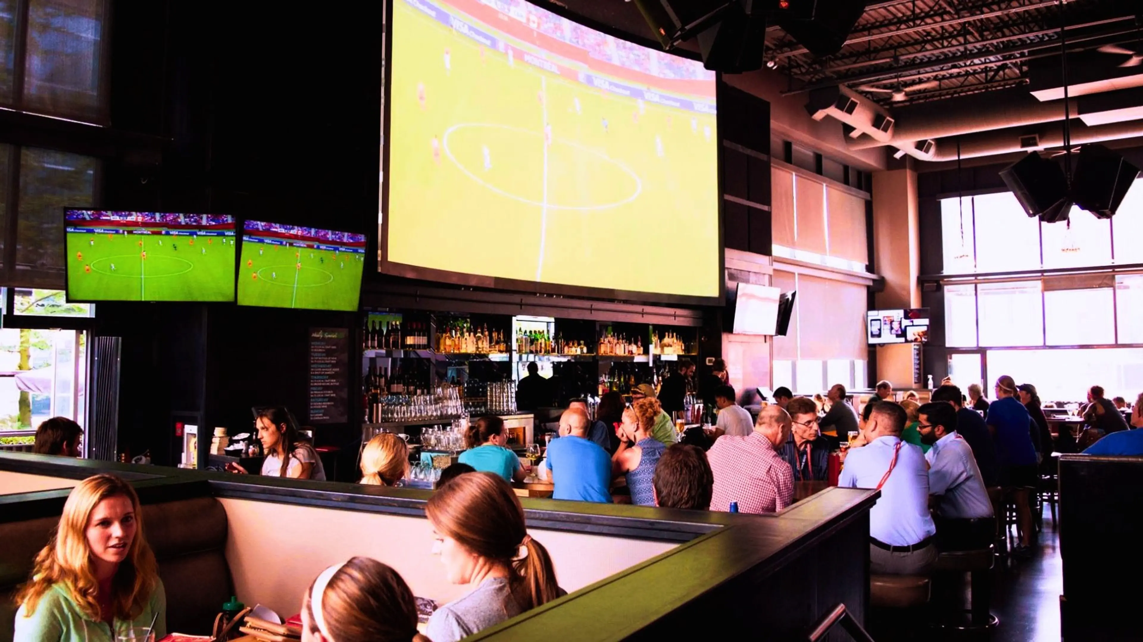 People sitting in a bar watching a soccer game on multiple large screens mounted on the walls.