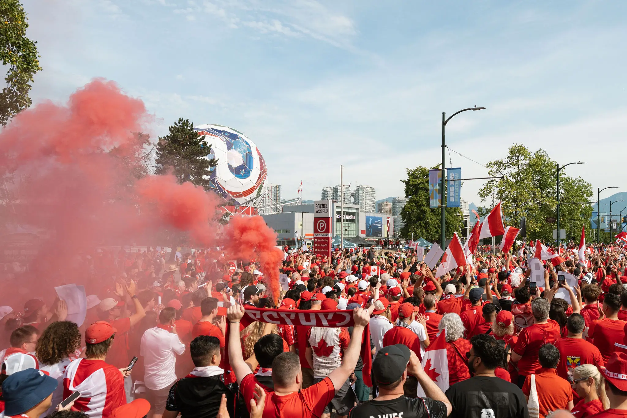 Large crowd wearing red and white, many holding Canadian flags and a red smoke flare in the air during an outdoor event.