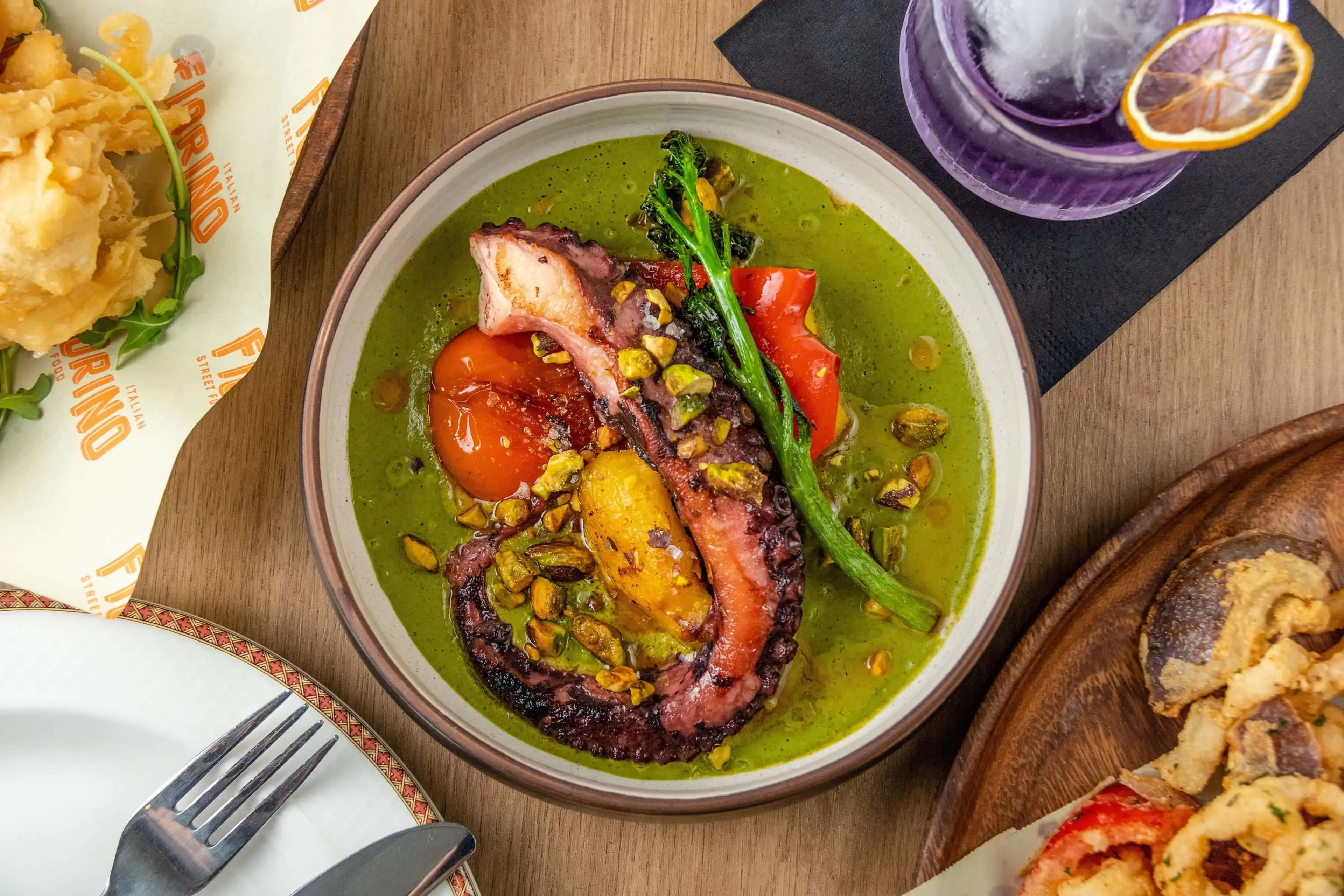 A vibrant overhead shot of a dish with a large piece of grilled octopus in a green sauce, garnished with red pepper and broccolini. A drink with a lemon slice sits to the side, and a separate plate with fried items is partially visible.