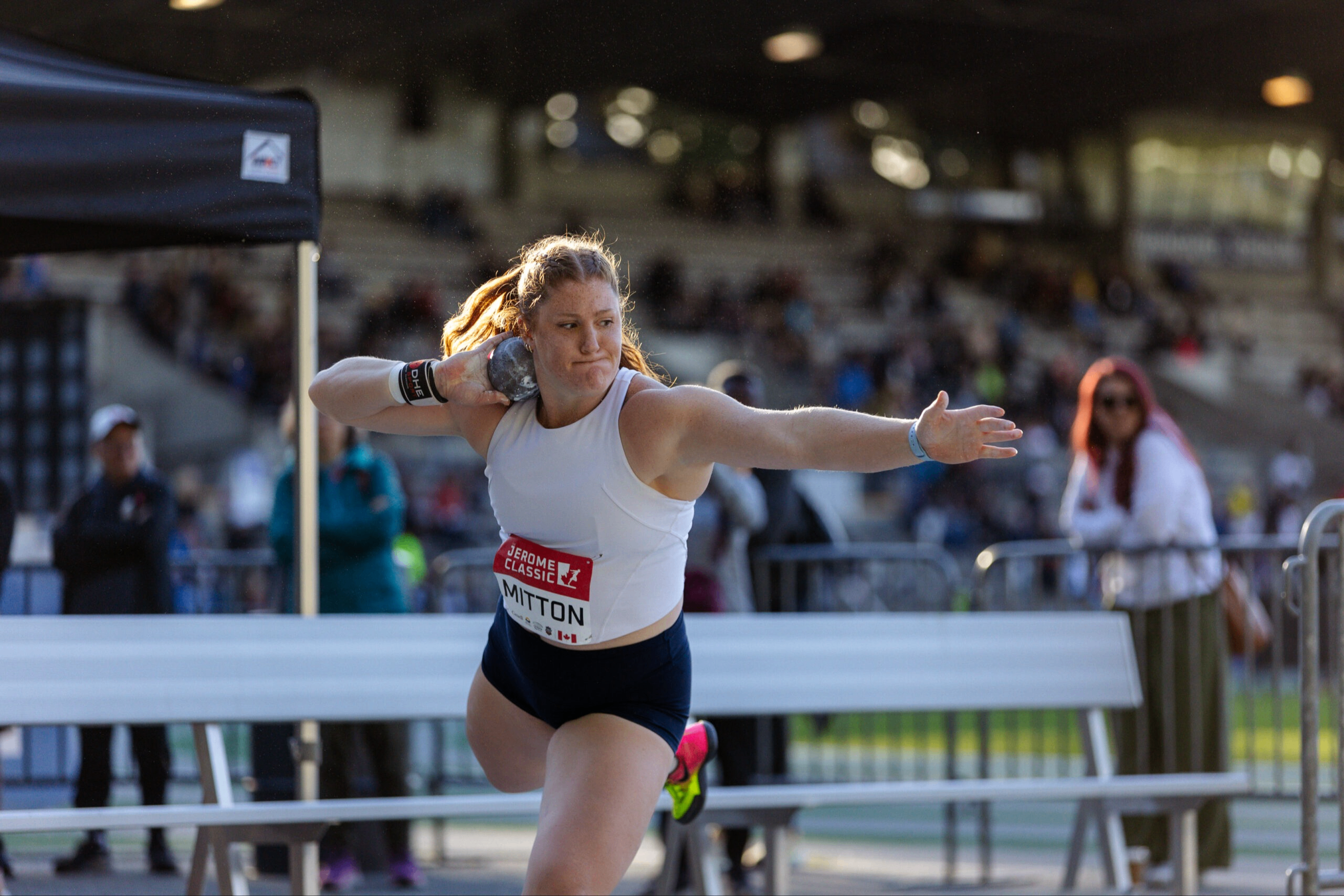 Athlete in white tank top and black shorts preparing to throw a shot put during a track and field event.