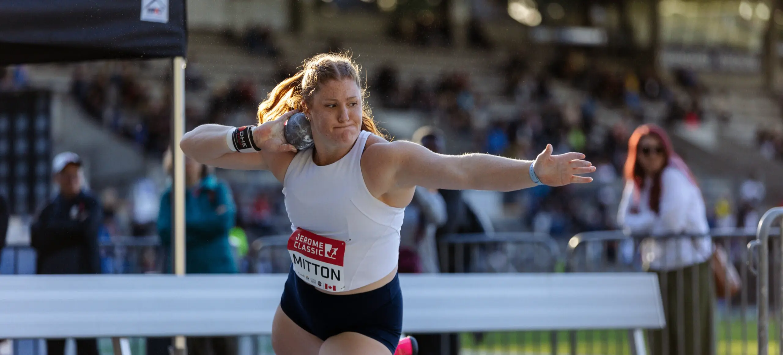 Athlete in white tank top and black shorts preparing to throw a shot put during a track and field event.