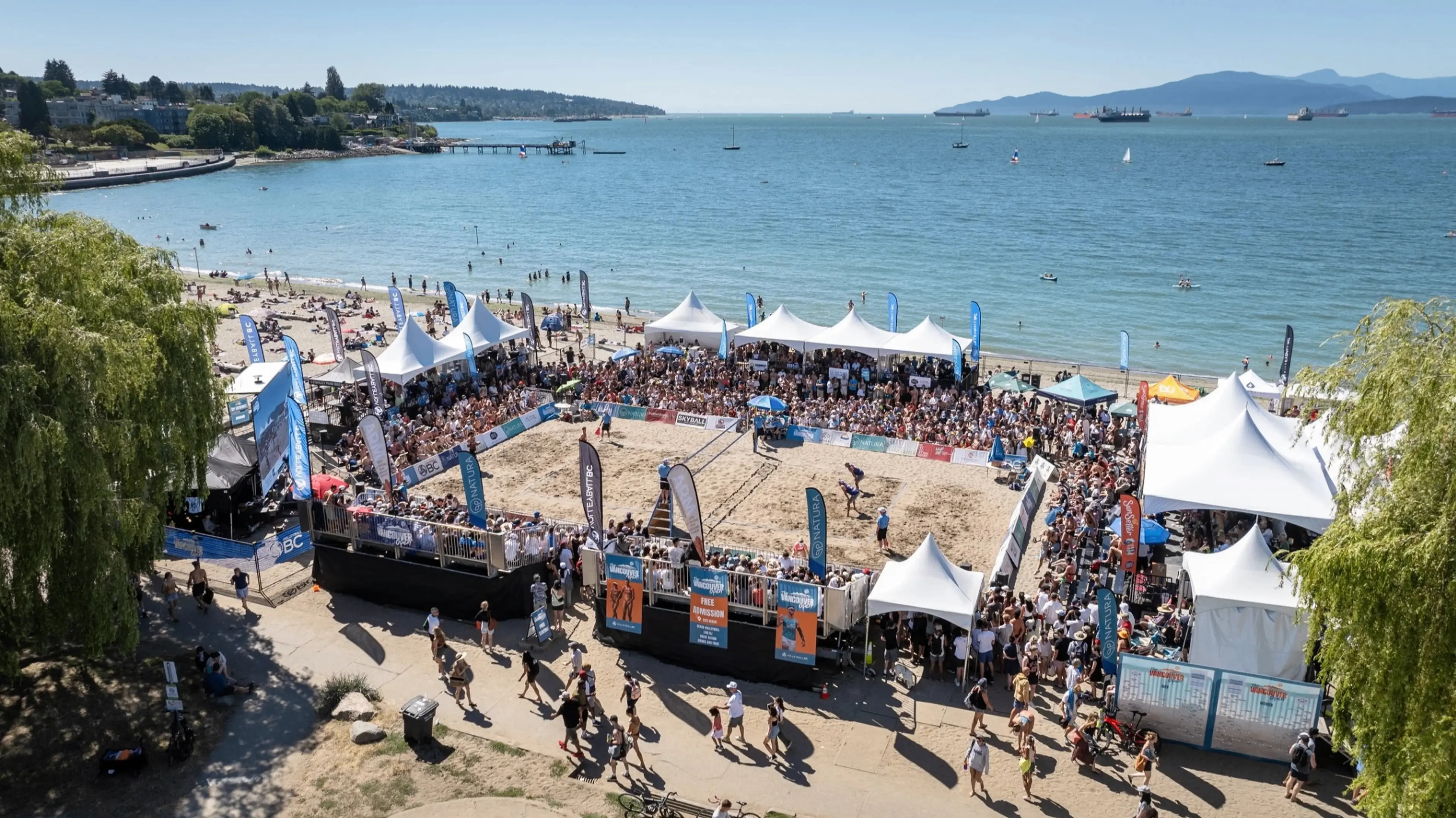 Crowded beach volleyball court with spectators under white tents by the ocean on a sunny day.
