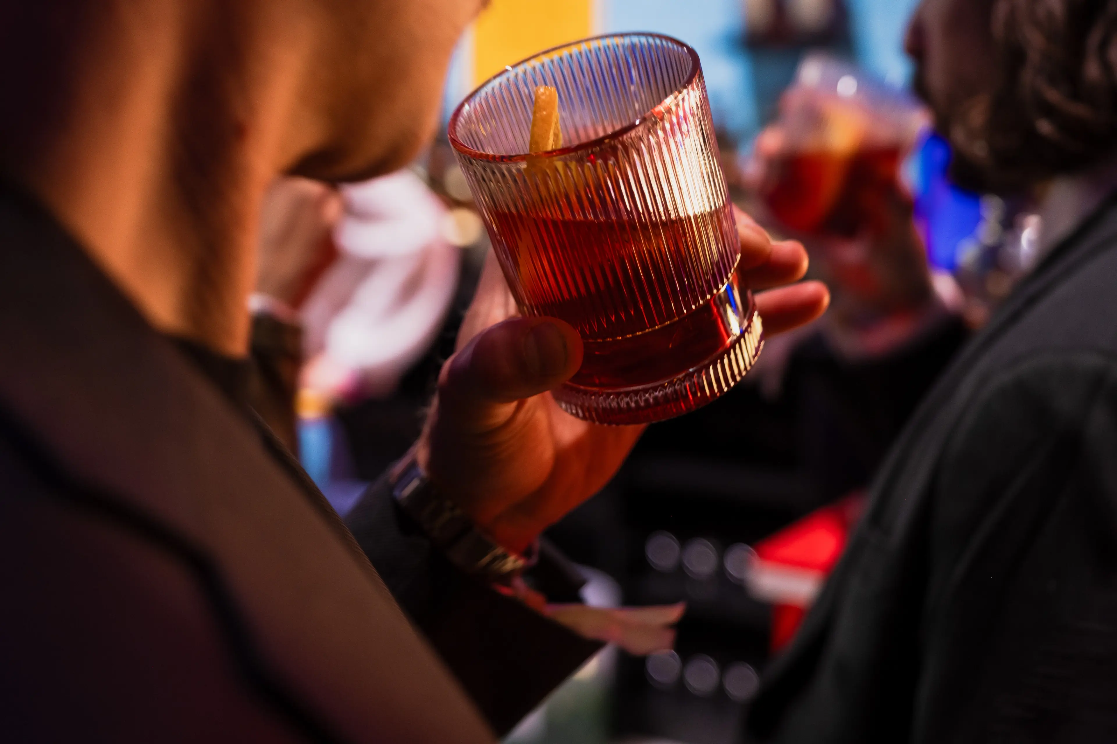 Person holding a glass of dark red drink with an orange peel garnish in a social setting.