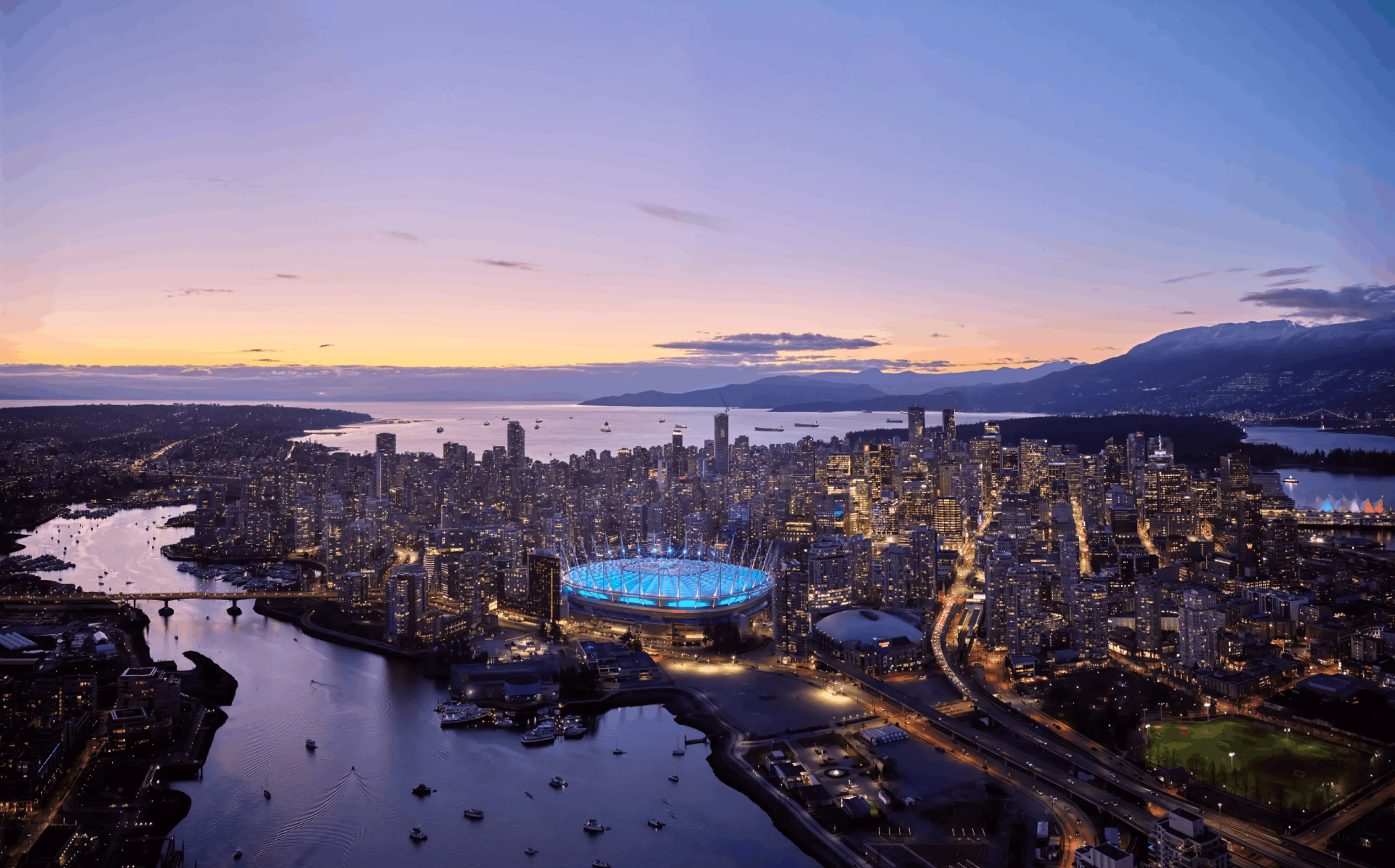 City skyline at dusk with a stadium lit in blue and its reflection in the water.