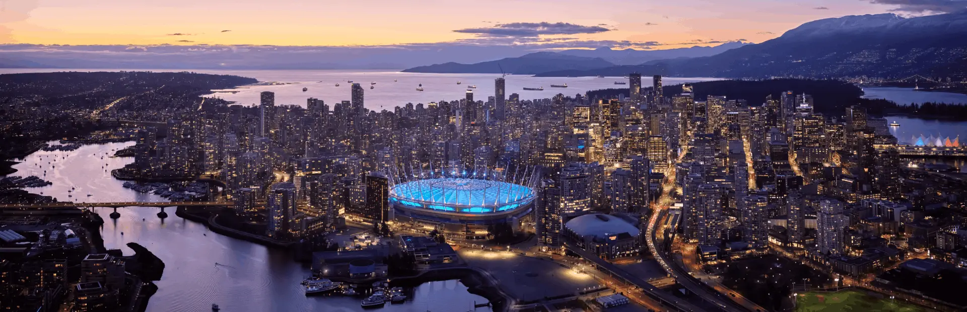 Aerial view of a city at dusk with a stadium lit in blue, surrounded by water and tall buildings under a purple and orange sky.