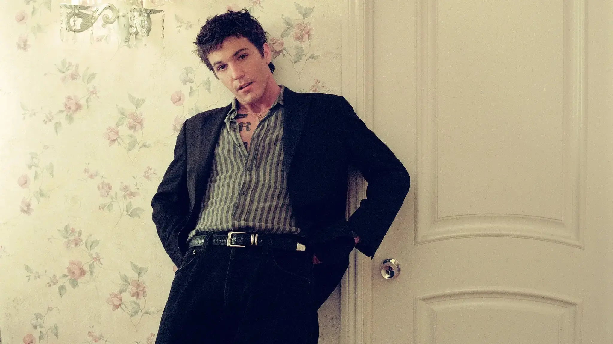 Person with short dark hair wearing a striped shirt and black jacket leaning against a floral wallpapered wall and a white door.