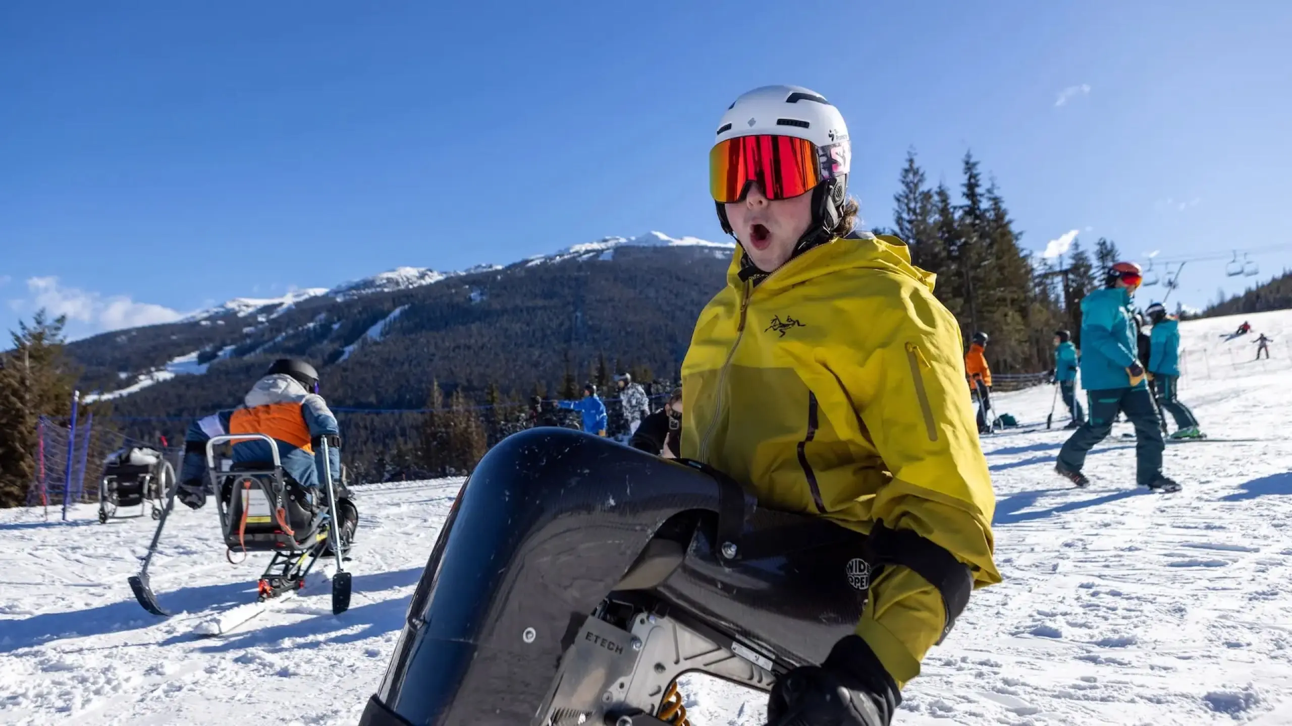 Person in yellow jacket and white helmet holding a sit-ski on a snowy mountain with other skiers and trees in the background.