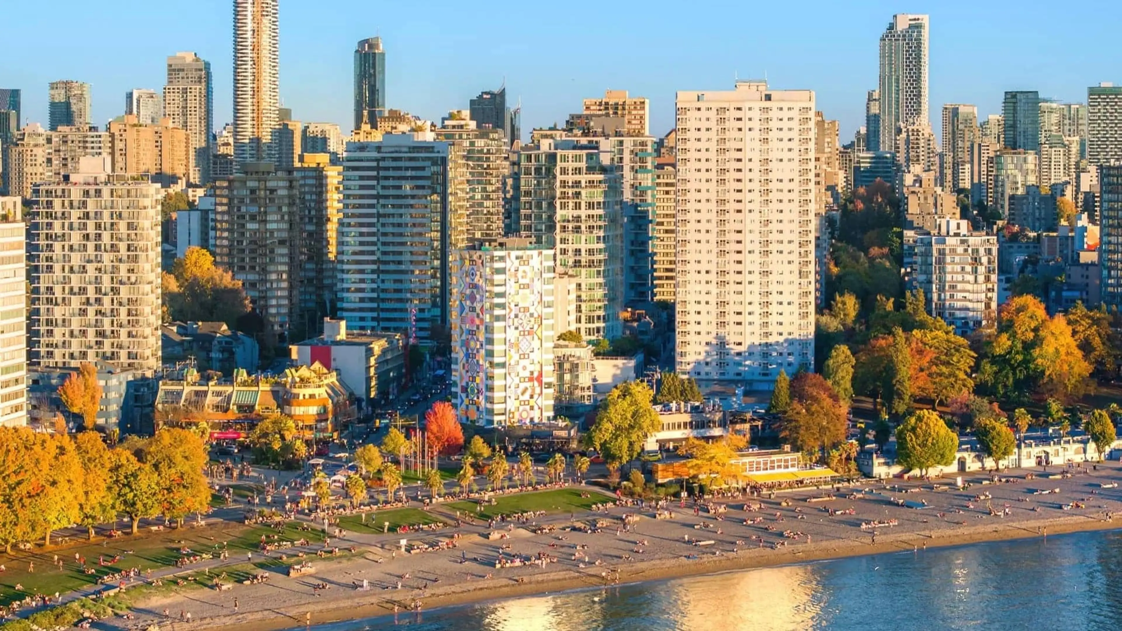 City skyline with tall buildings and a beach with people along the waterfront on a sunny day.
