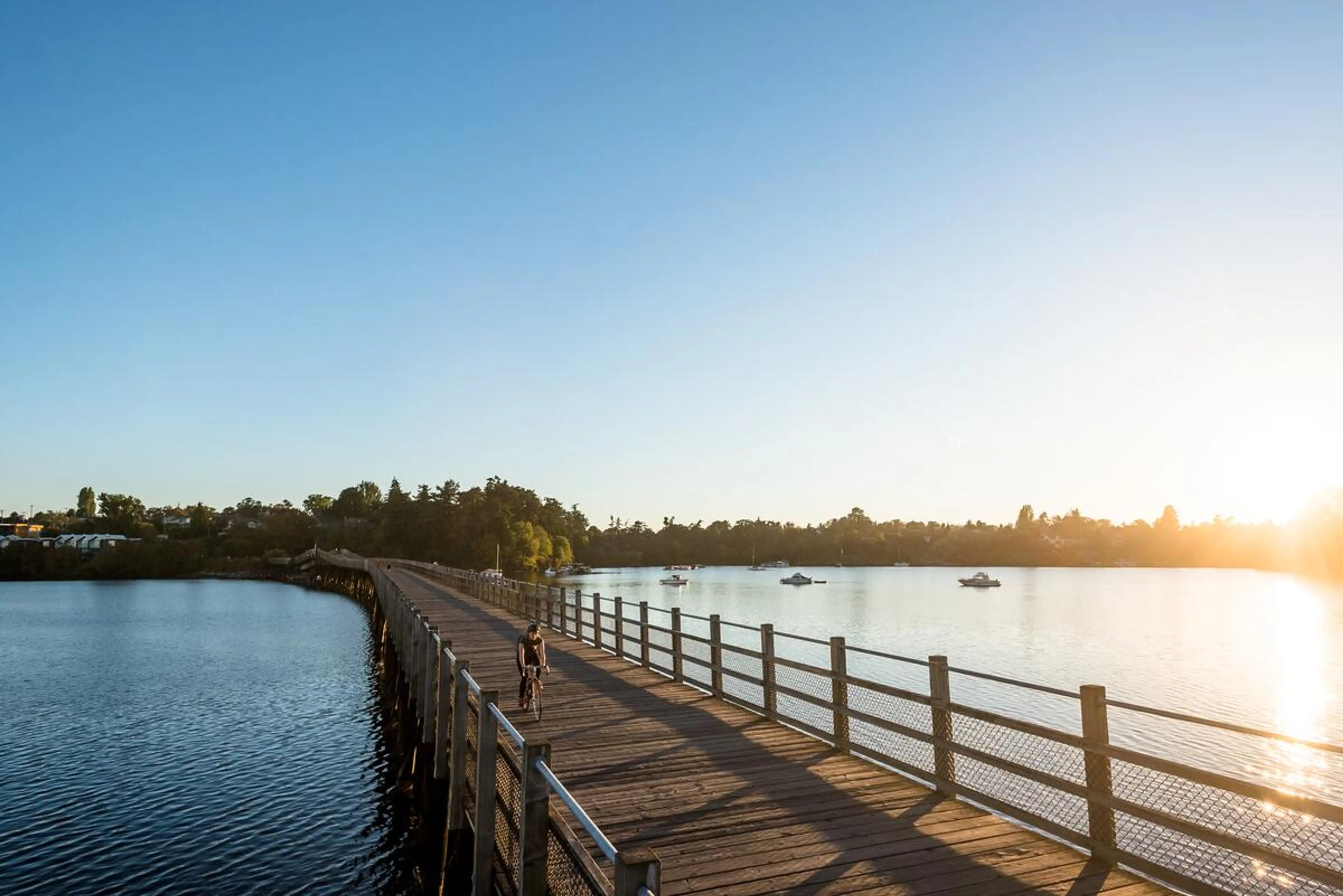 Person riding a bicycle on a wooden bridge over water during sunset with boats in the distance.