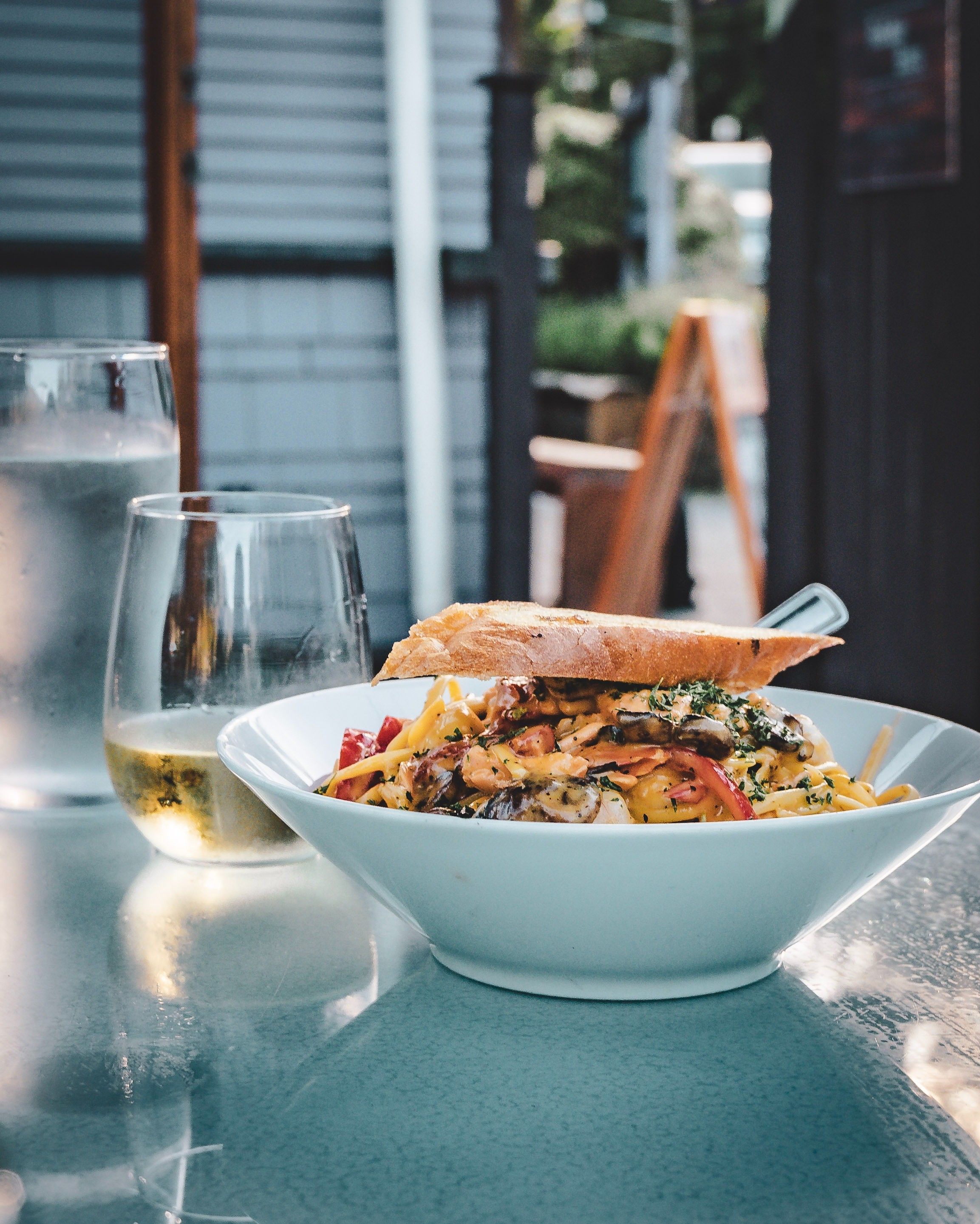 Bowl of pasta with vegetables and herbs topped with a slice of bread on a table next to a glass of white wine and a glass of water.