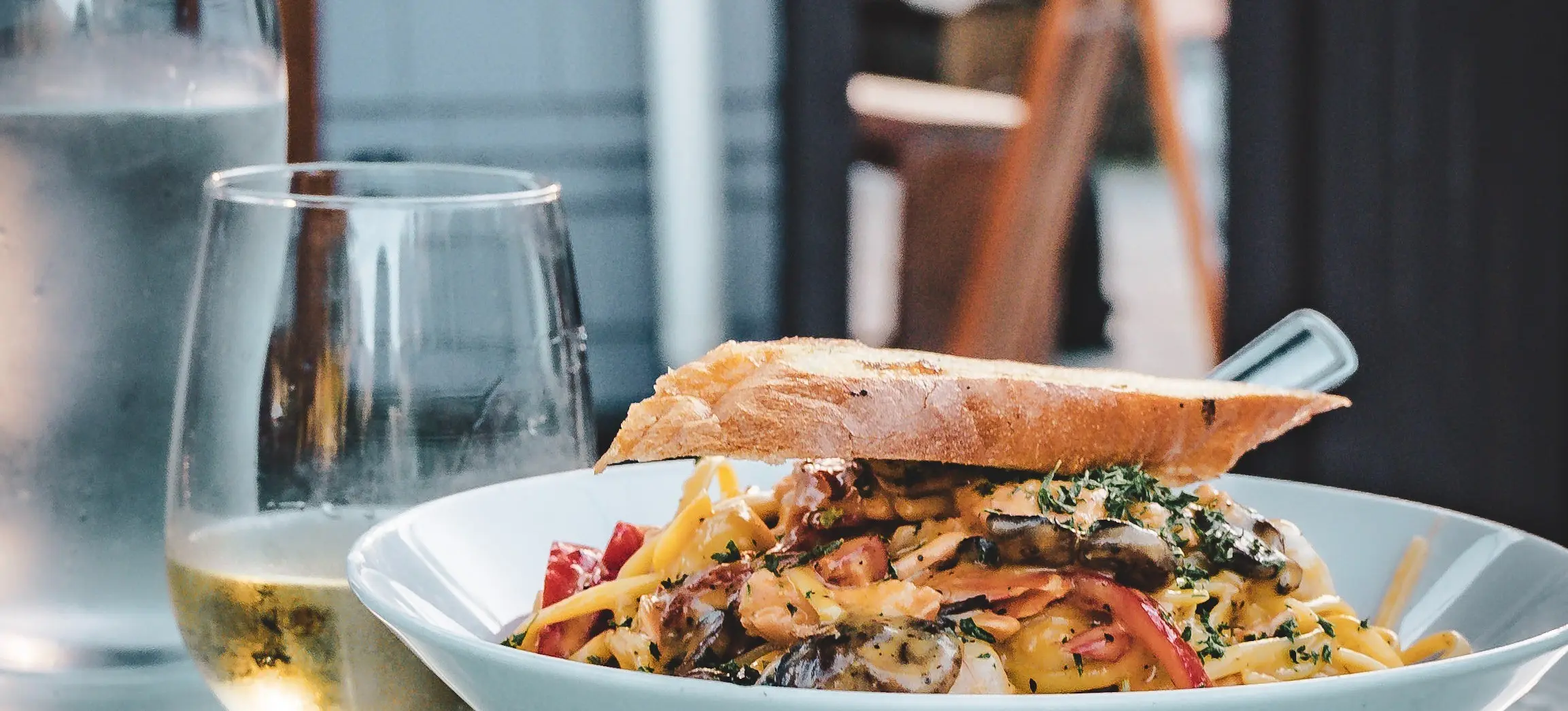 Bowl of pasta with vegetables and herbs topped with a slice of bread on a table next to a glass of white wine and a glass of water.