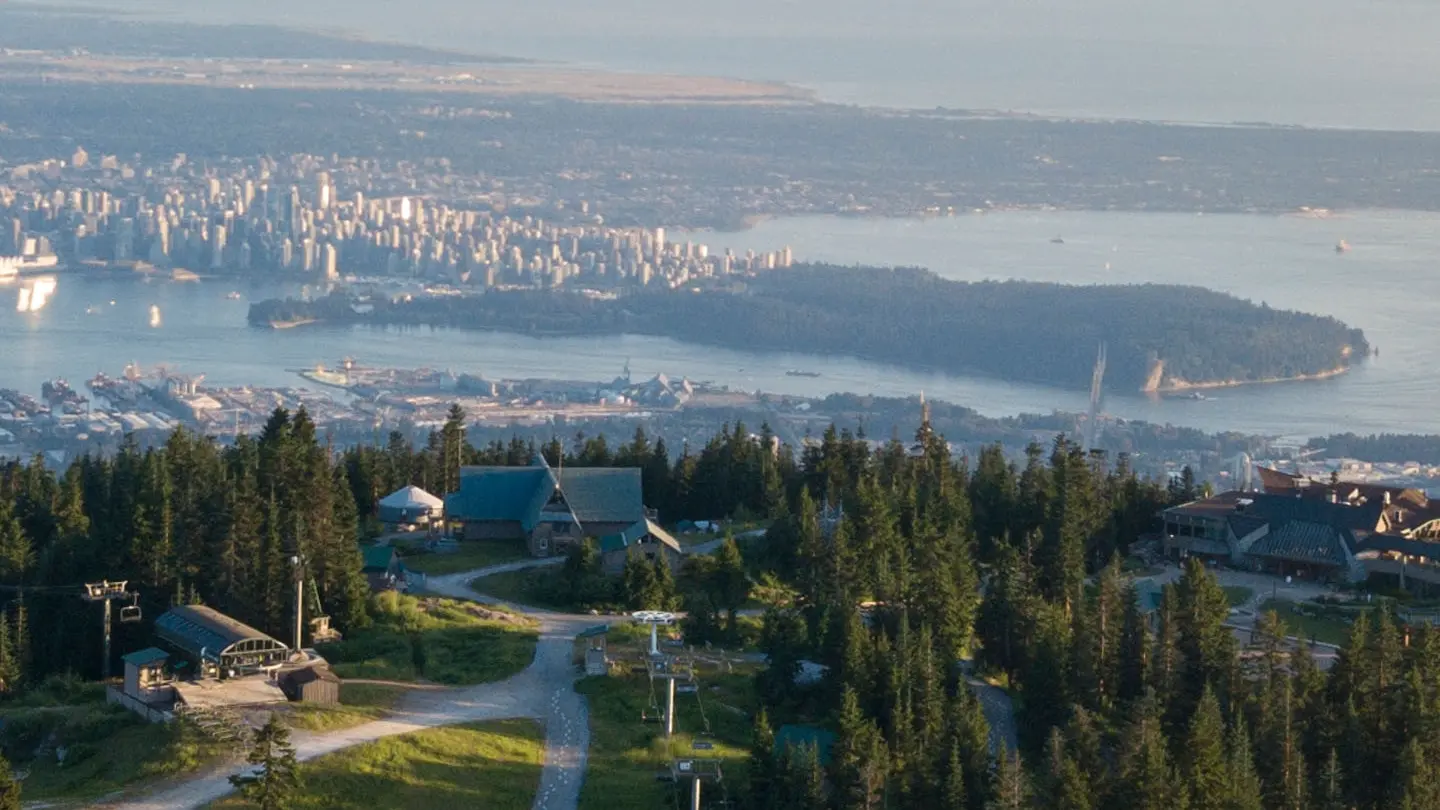 View of a forested mountain area with buildings and ski lifts overlooking a city and water body in the distance.