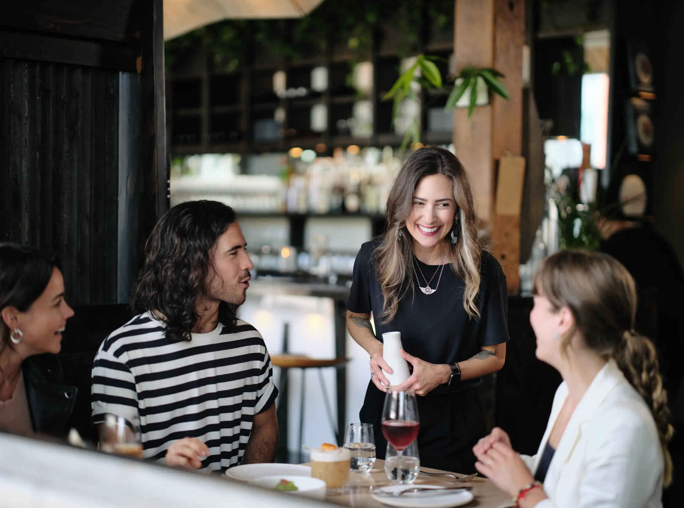 A person holding a white bottle smiles and talks to three people seated at a restaurant table with drinks and plates.