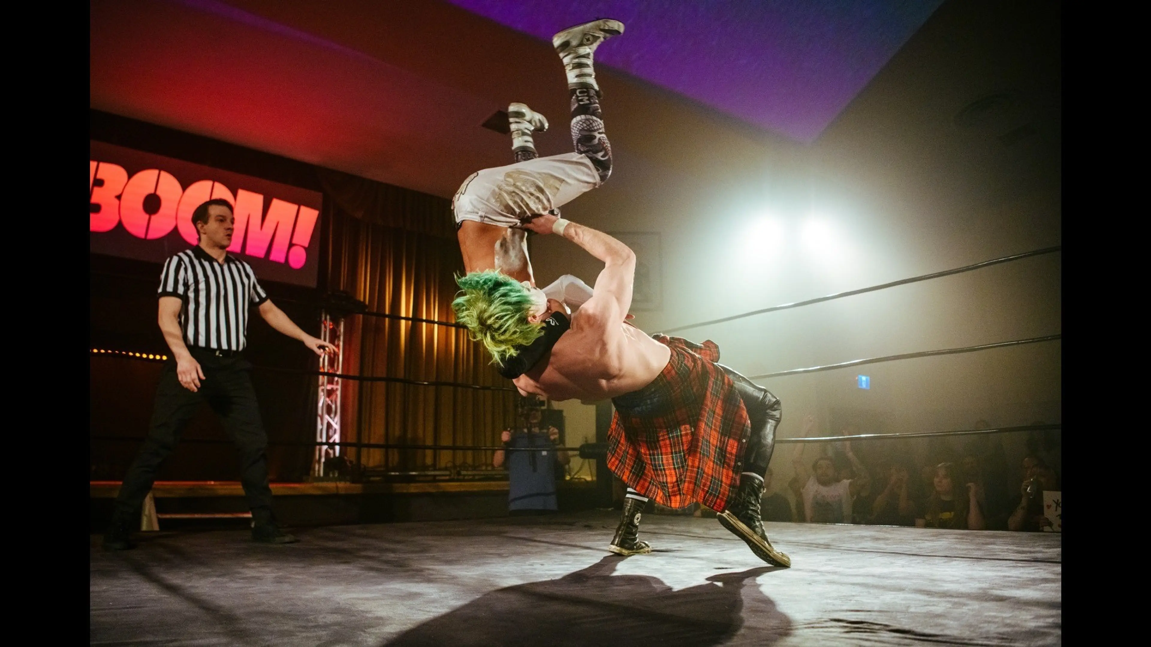 Two wrestlers in a ring performing a move with one lifting the other upside down, a referee watches nearby.