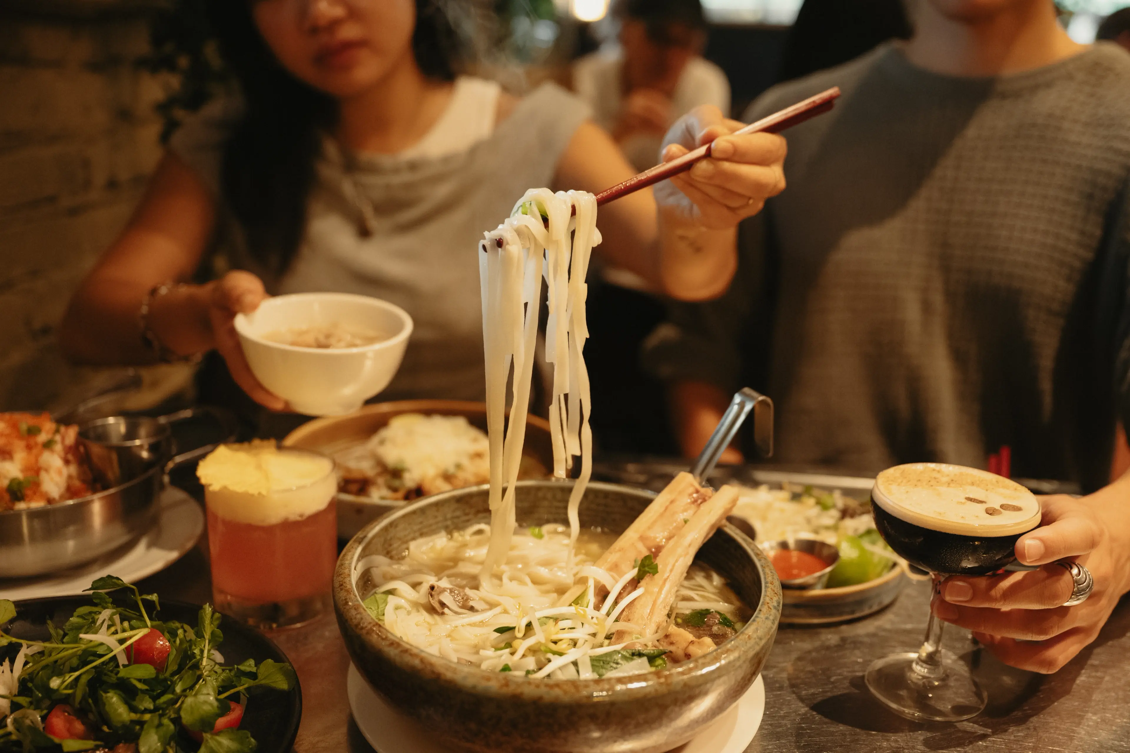 Person lifting noodles with chopsticks from a bowl of soup with ribs, another person holding a dark drink in a glass, and various dishes on the table.