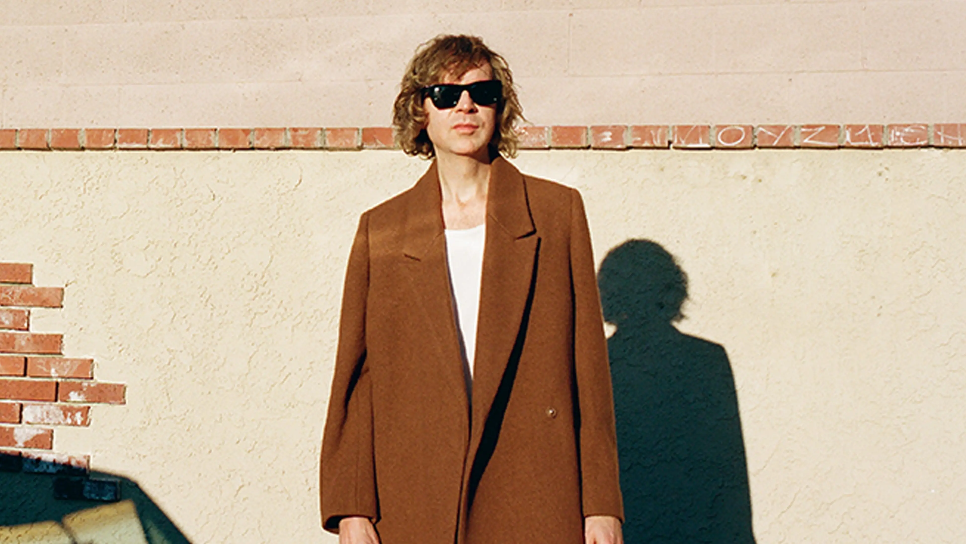 Person wearing a brown coat and black sunglasses standing against a beige wall with a shadow cast on it.