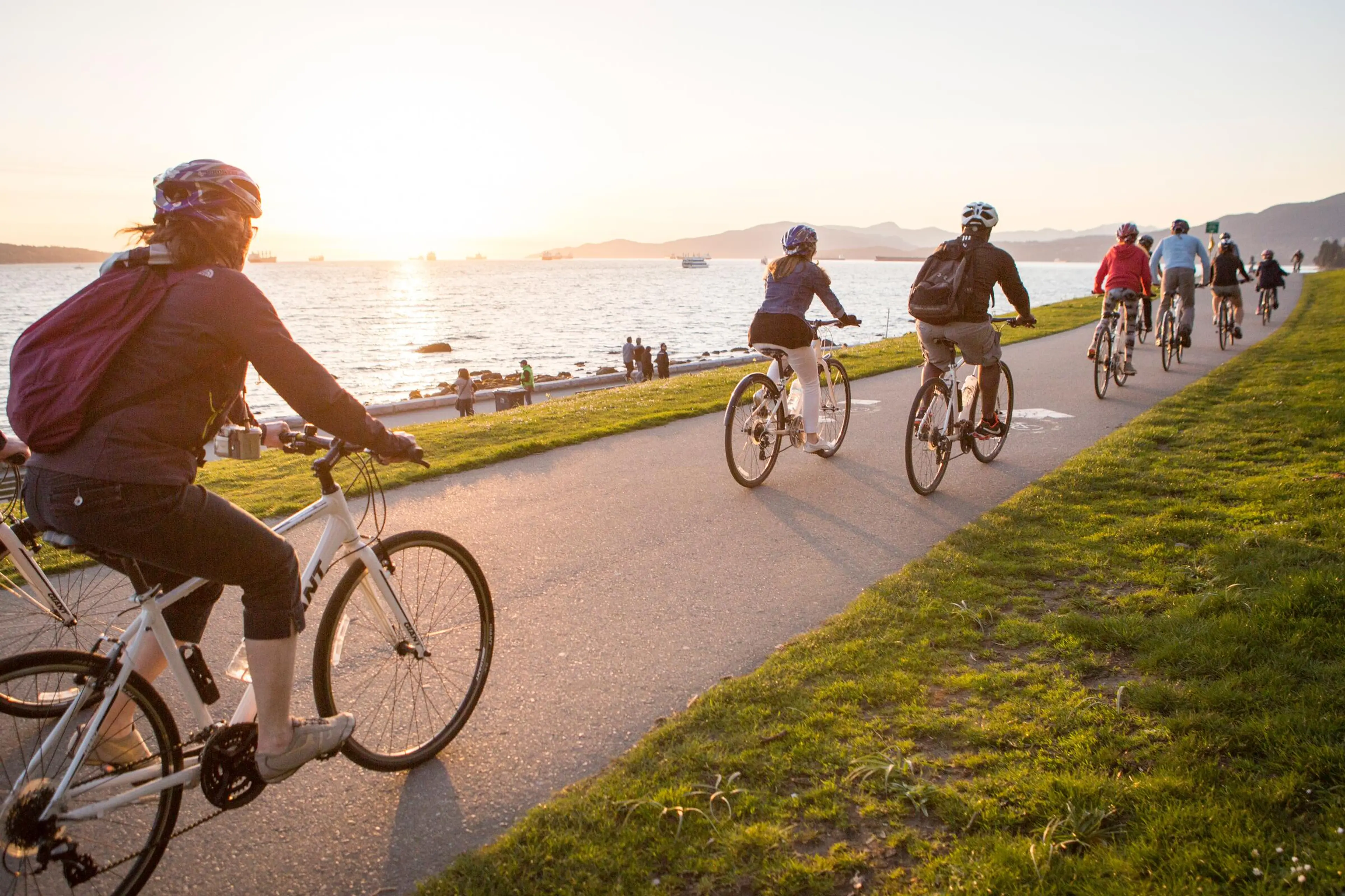 Group of people cycling on a paved path along a waterfront at sunset with mountains in the background.