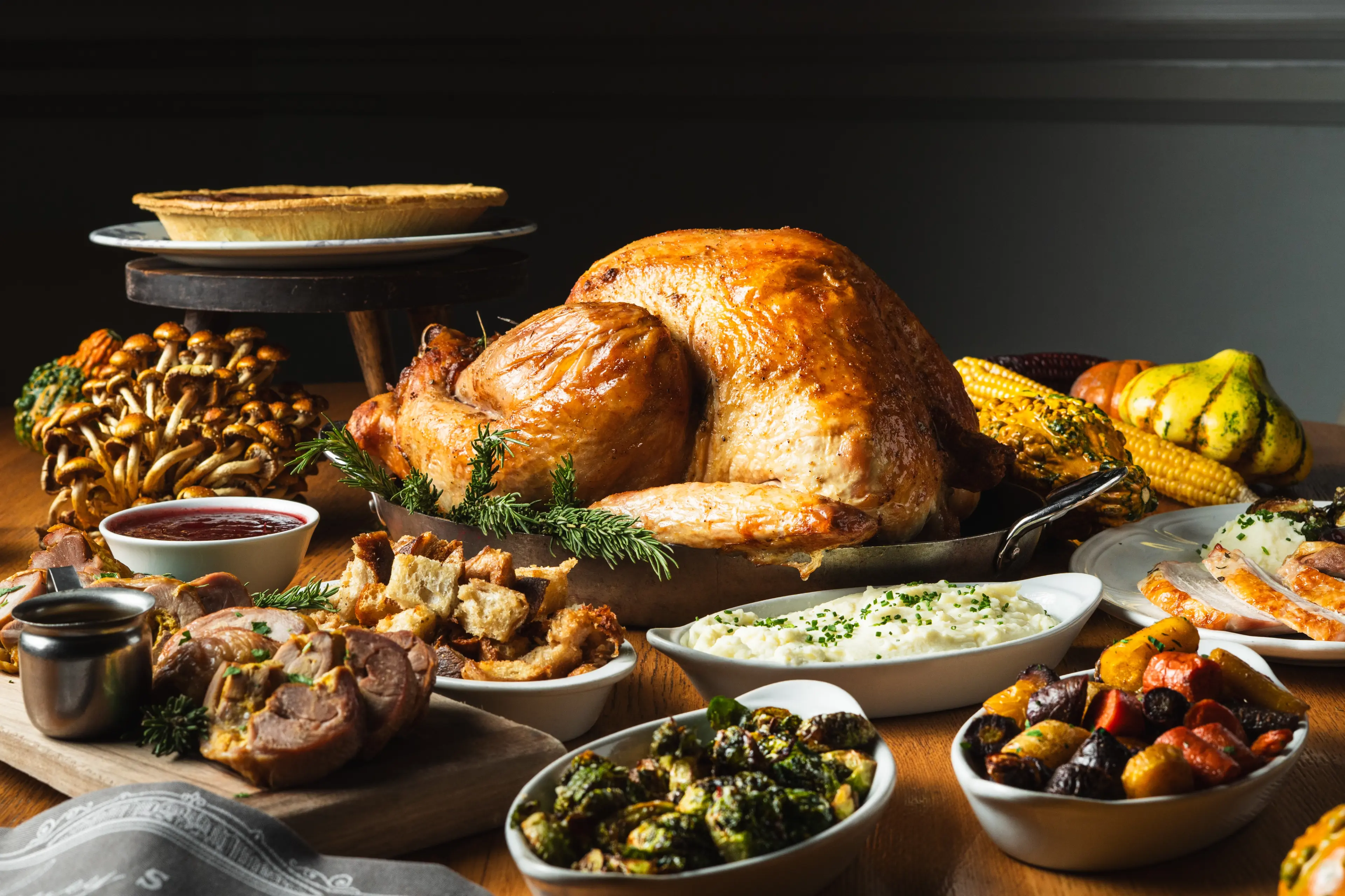 Roasted whole turkey surrounded by mashed potatoes, stuffing, roasted vegetables, cranberry sauce, pie, and corn on a wooden table.