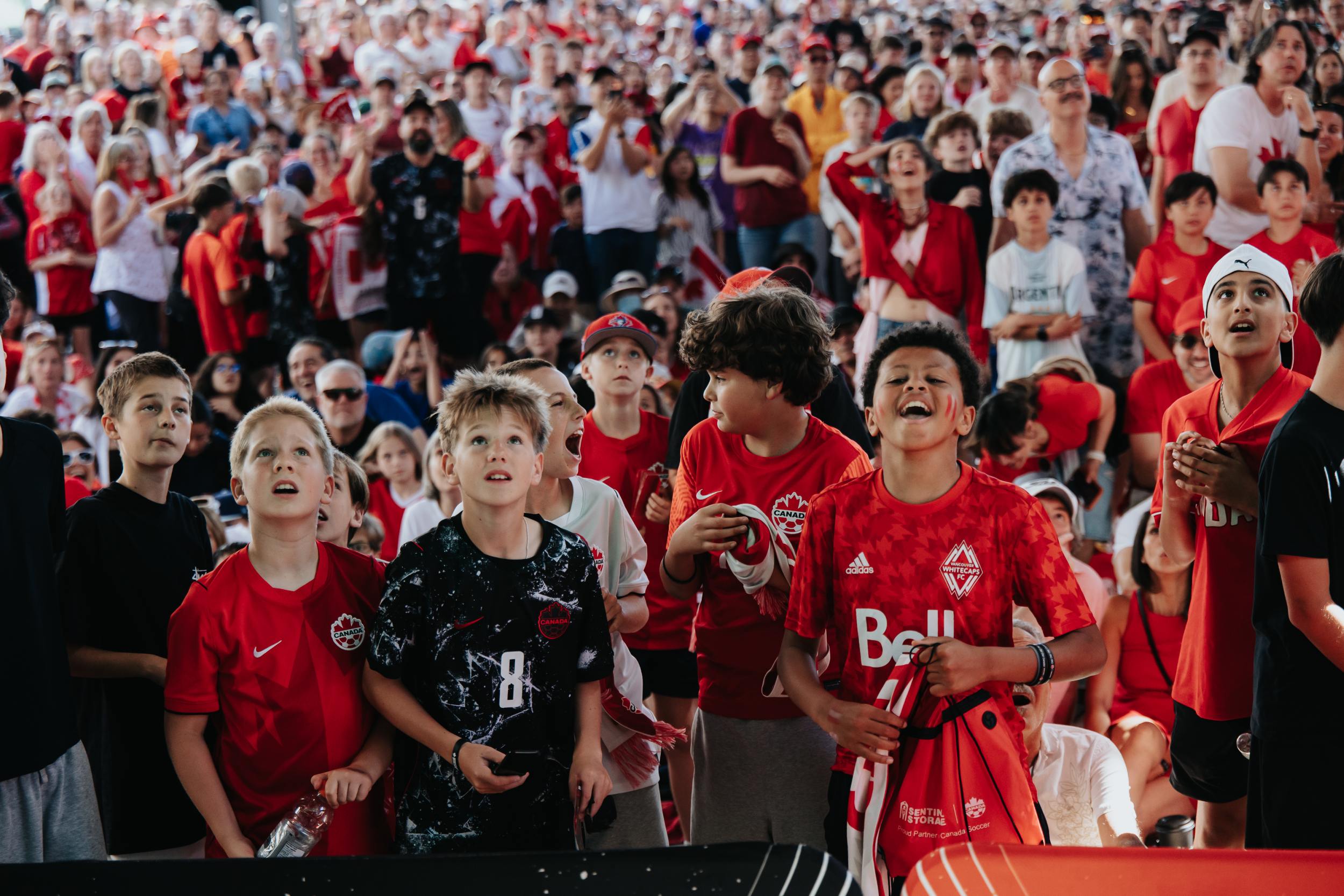 Group of children and adults in red and black sportswear watching an event, some cheering and looking excited.