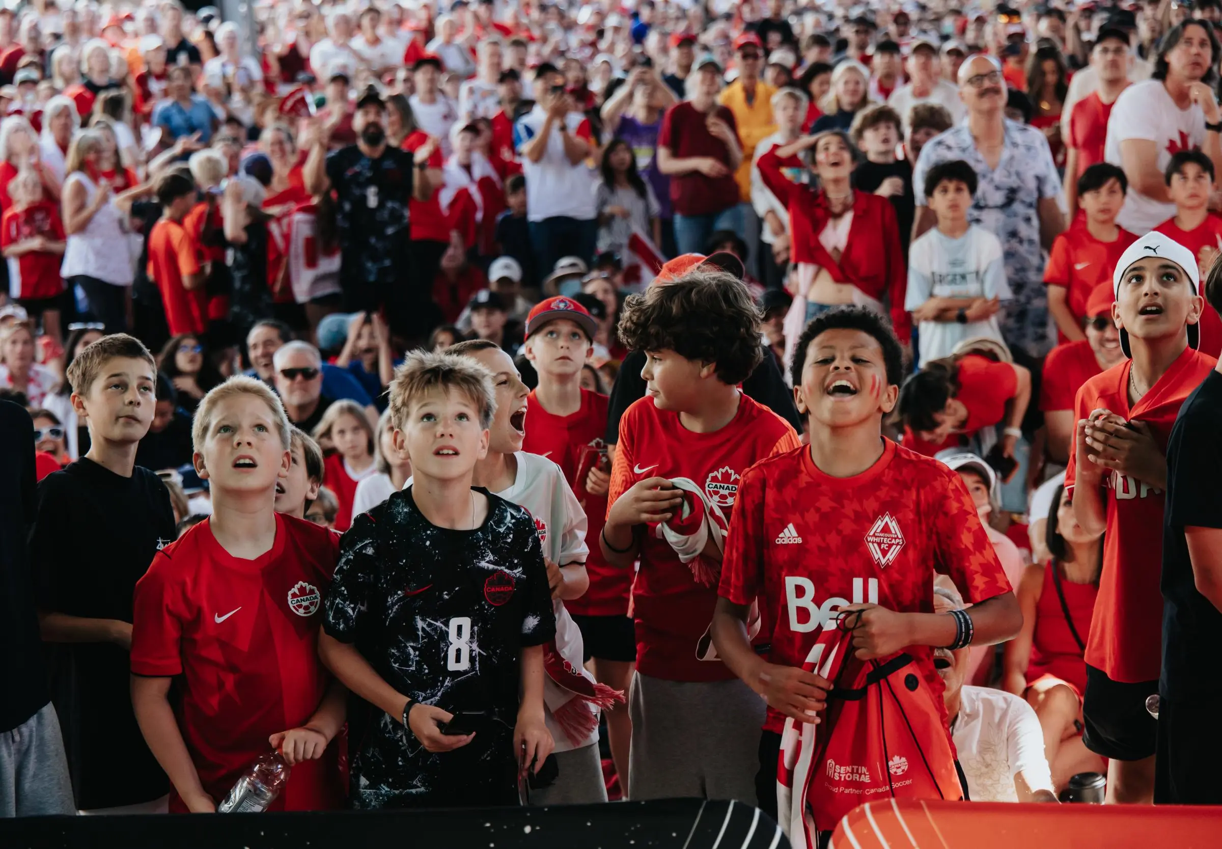 Group of children and adults in red and black sportswear watching an event, some cheering and looking excited.