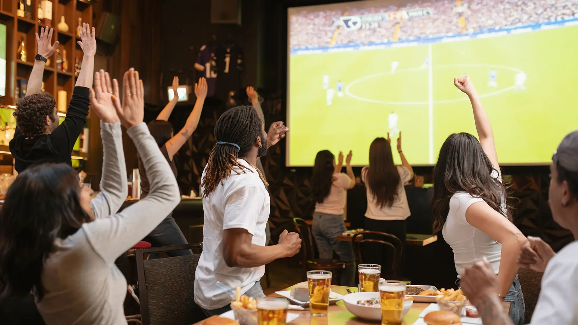 A group of people in a bar cheering and raising hands while watching a soccer game on a large screen.
