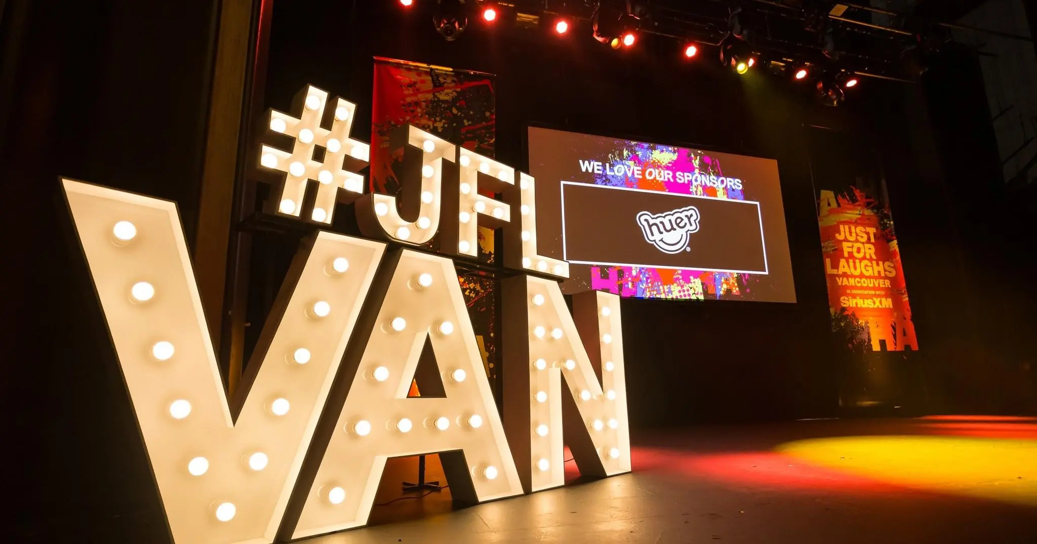 Large illuminated letters spelling #JFL VAN on a stage with colorful sponsor signage in the background.
