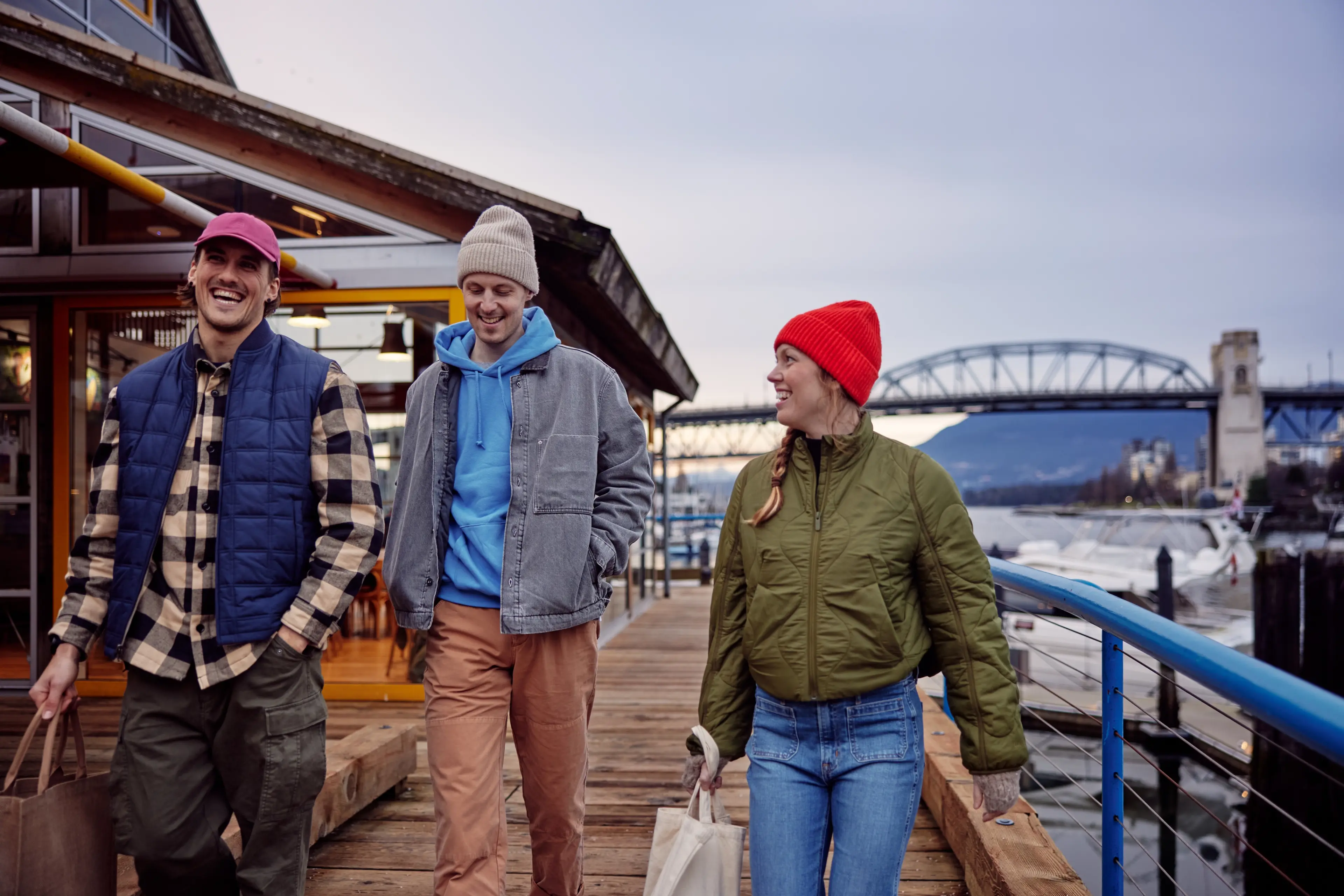 A group of friends walking on a wooden dock near water on Granville Island, dressed in casual jackets and hats, smiling and carrying bags.