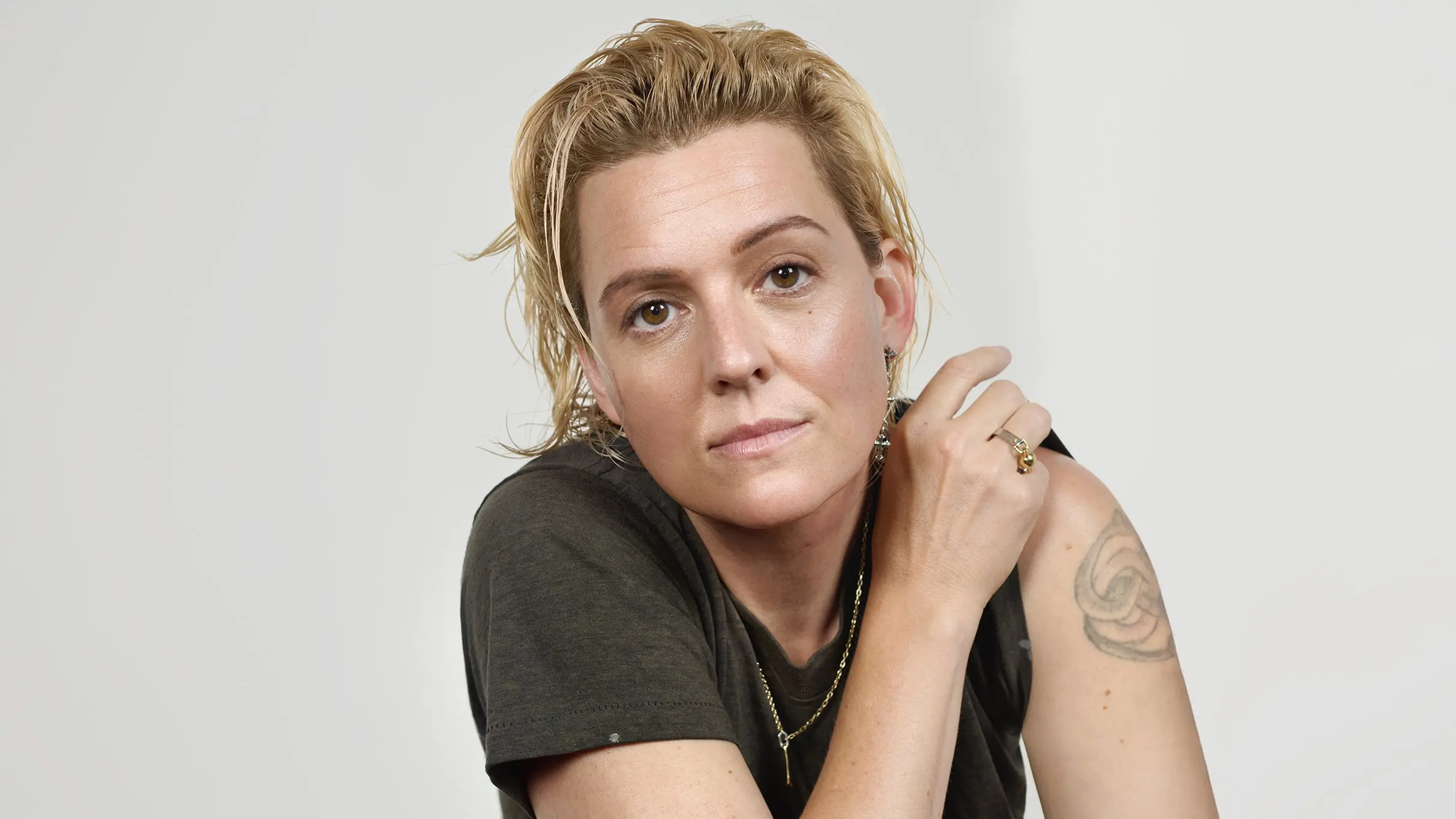 Person with short blonde hair wearing a dark shirt, showing a tattoo on their upper arm against a plain light background.