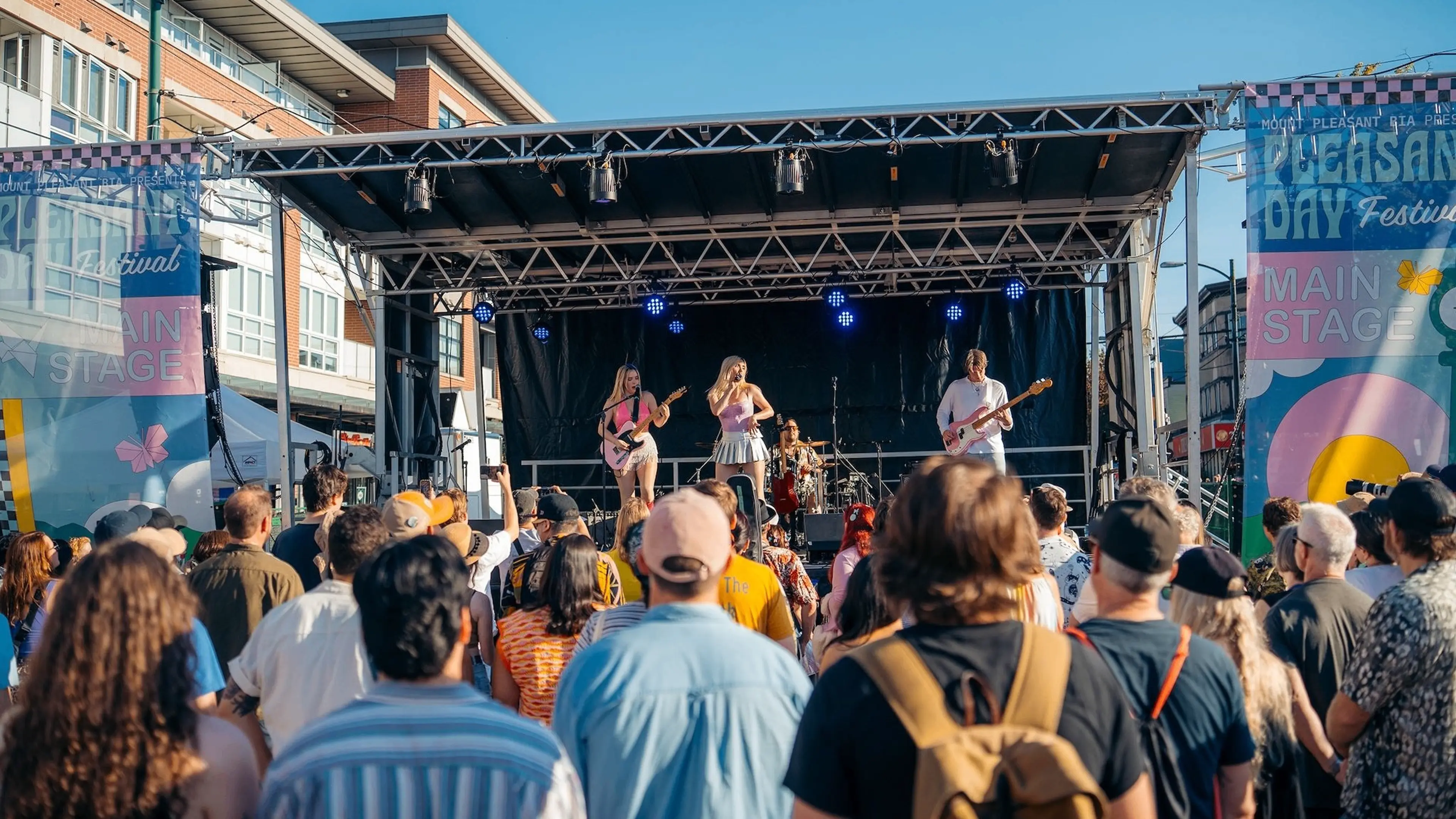 Crowd watching a band performing on an outdoor stage at a sunny festival with banners reading Pleasant Day Festival Main Stage.