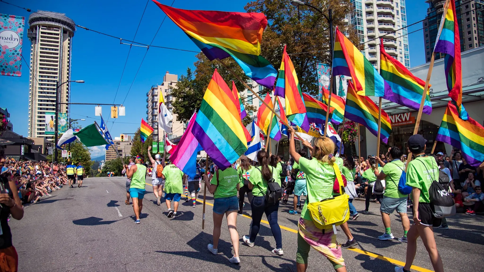 Group of people in green shirts marching on a city street waving rainbow pride flags during a parade.