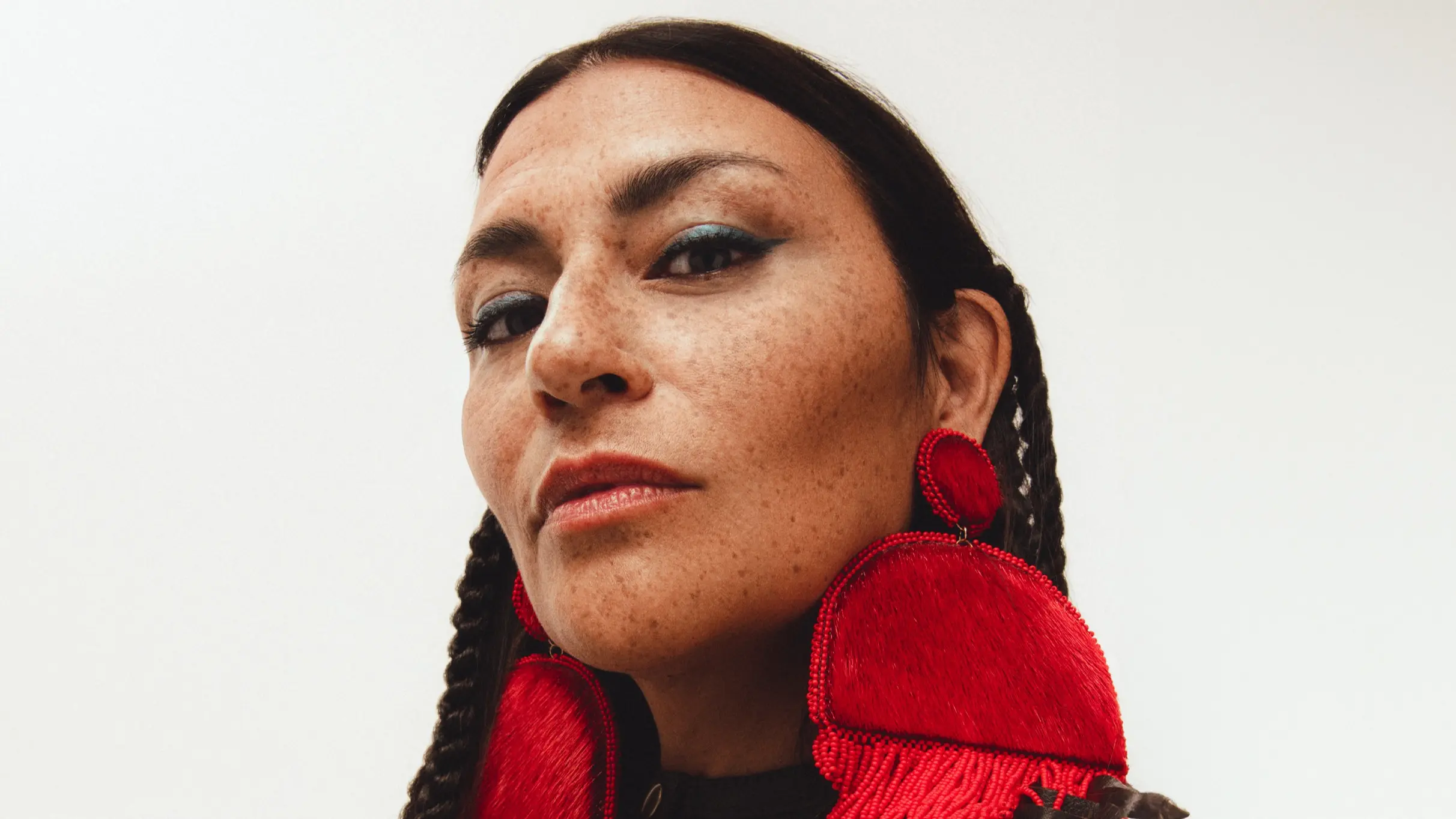 Close-up of a person with braided hair wearing large red earrings and subtle makeup against a white background.