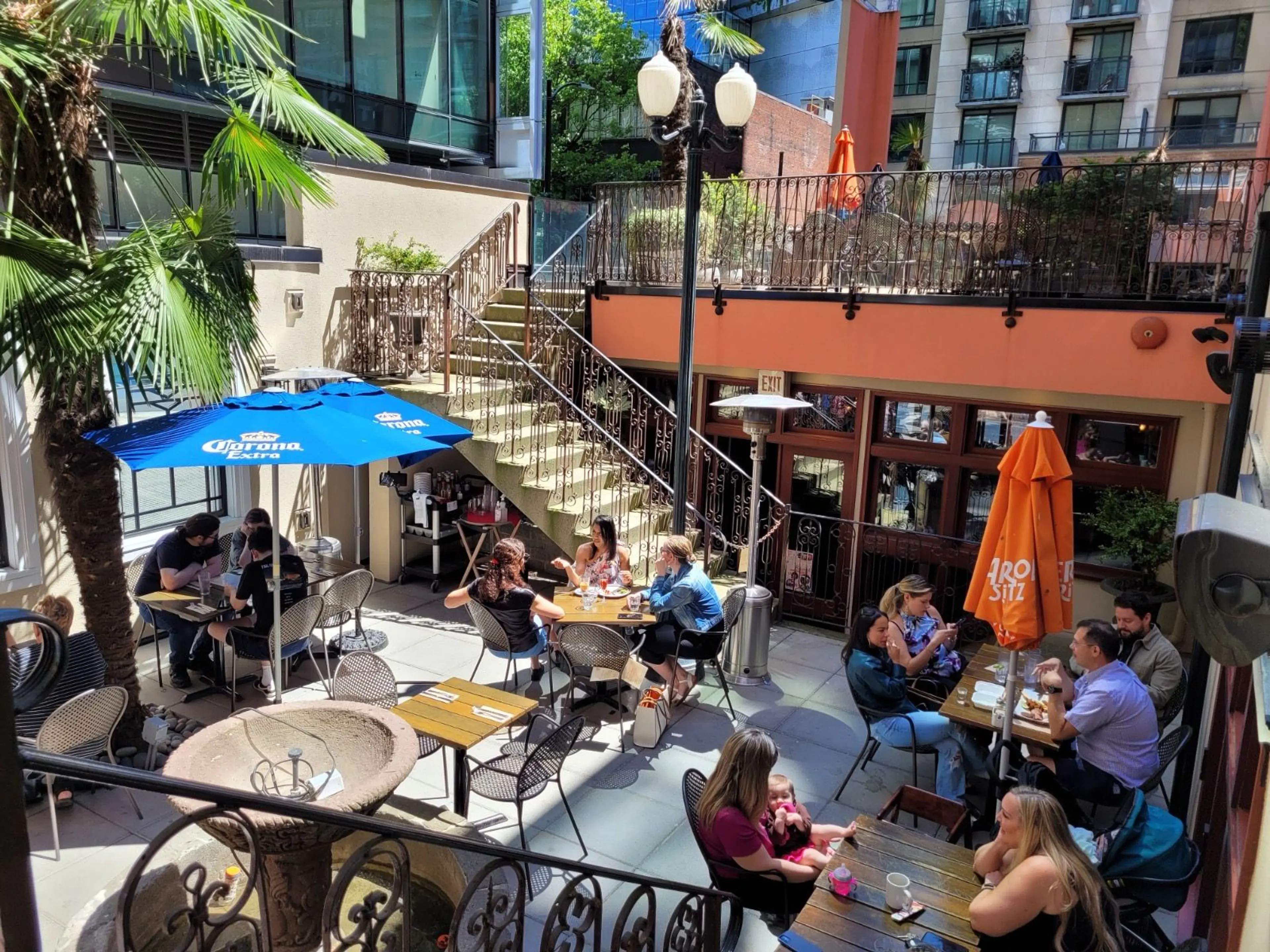Outdoor patio with people sitting at tables under umbrellas, surrounded by palm trees and stairs leading to an upper level.