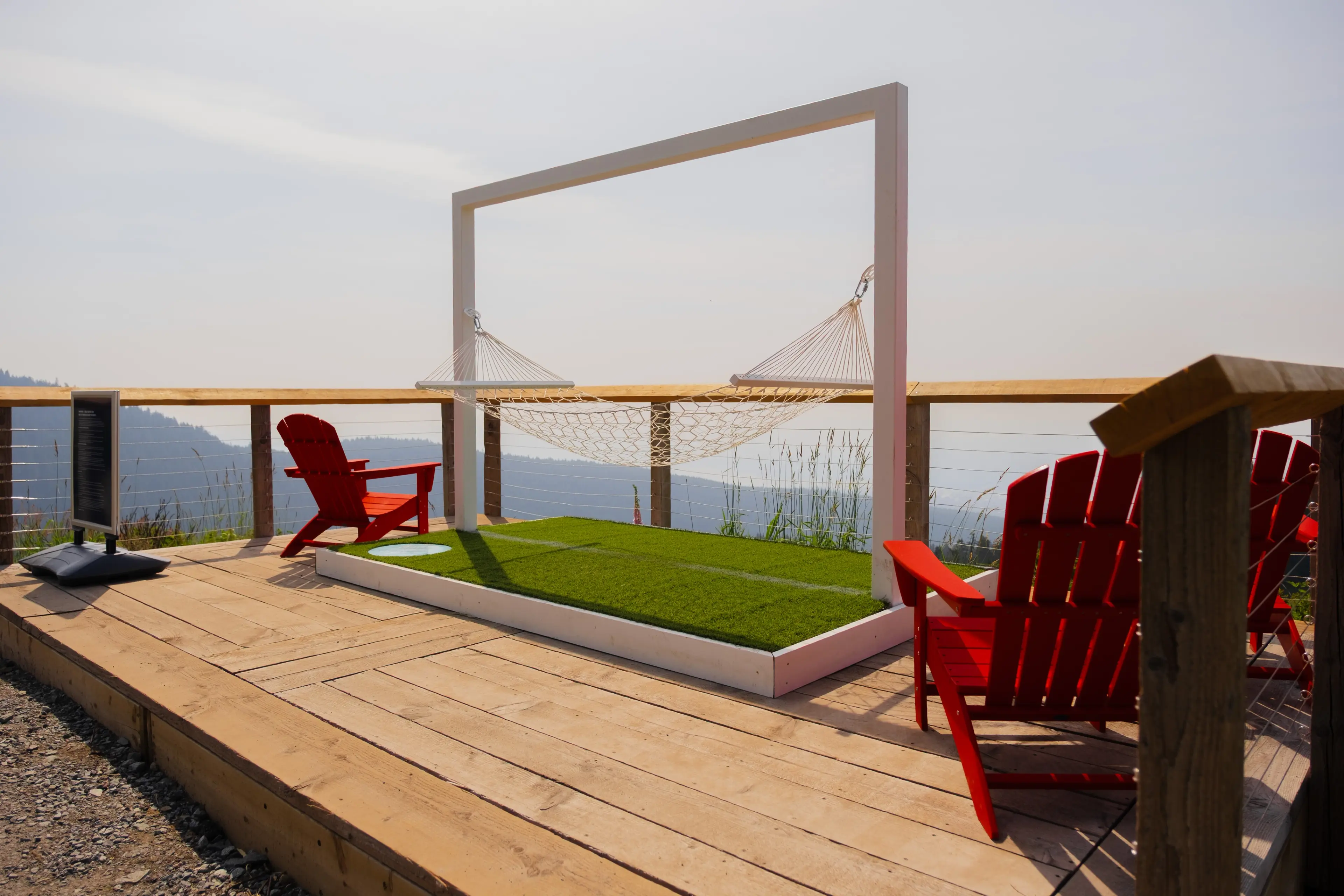 Outdoor wooden deck with red Adirondack chairs, a white hammock frame with a net hammock, and a grassy platform overlooking a mountainous landscape.