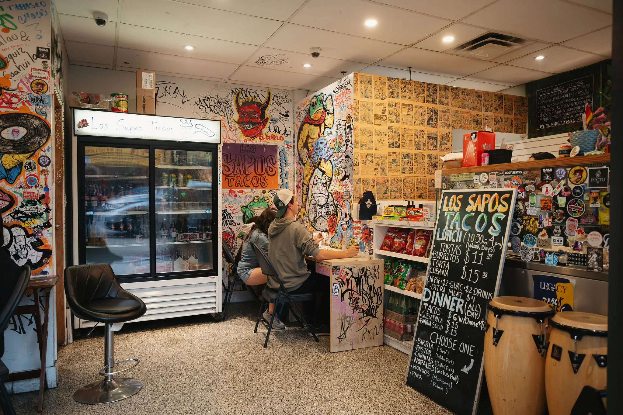 Two people sitting at a graffiti-covered counter in a colorful taco shop with a menu board listing tacos and prices, and a refrigerator on the left.