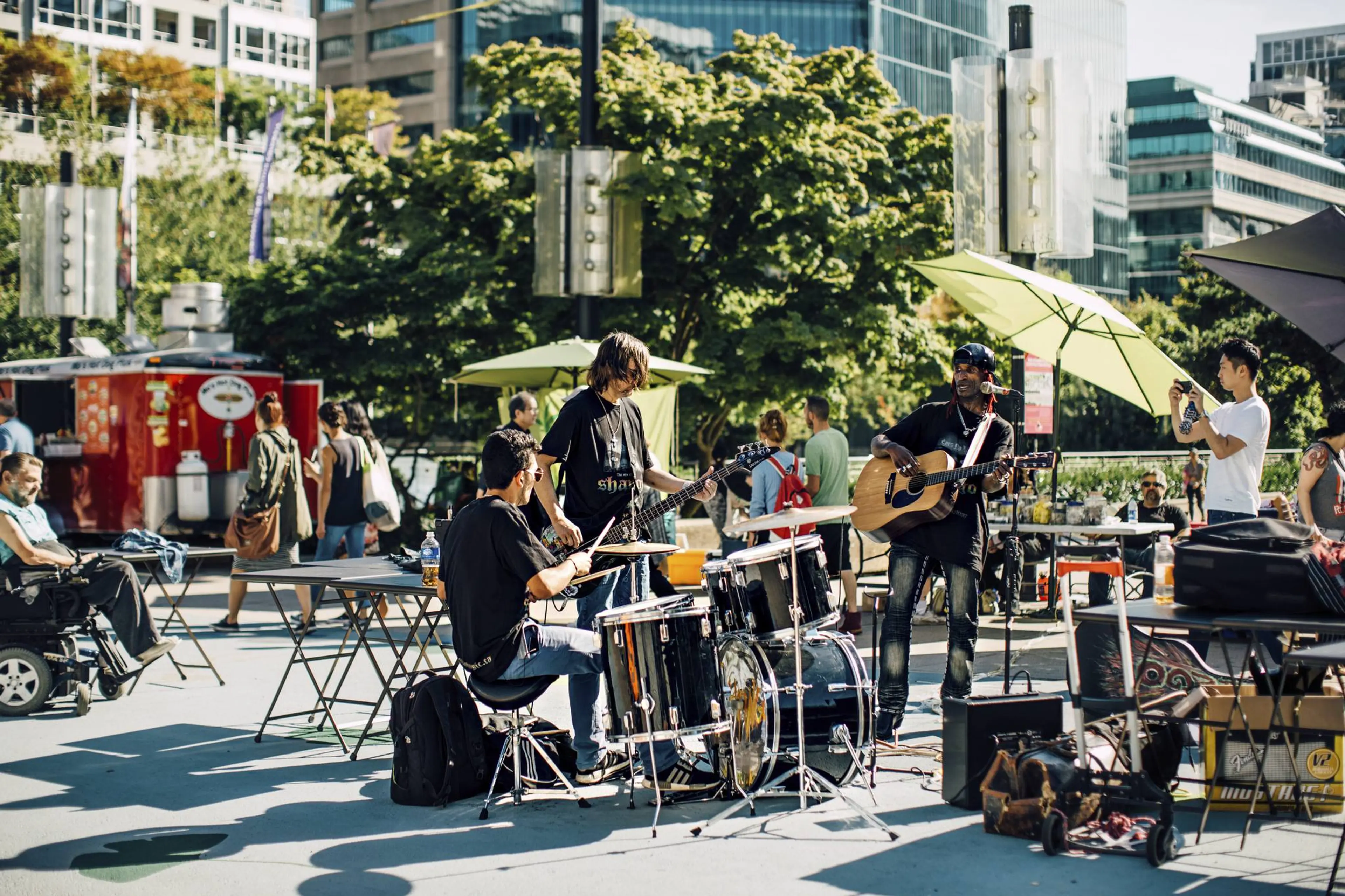 Outdoor scene with a three-person band playing drums, bass guitar, and acoustic guitar in a city plaza with people walking and sitting nearby.