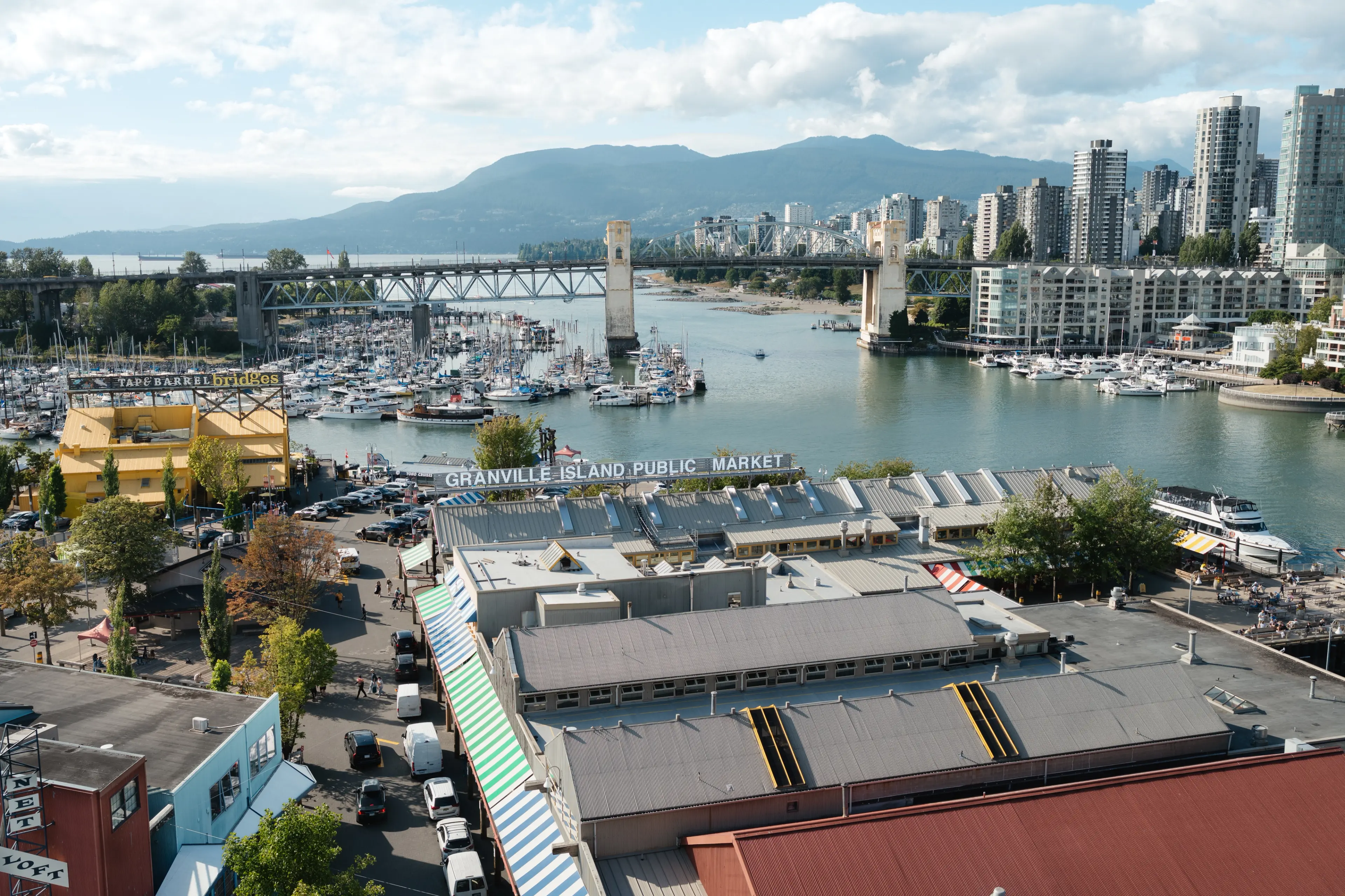 View of Granville Island Public Market, marina with boats, a bridge, and city buildings with mountains in the background.