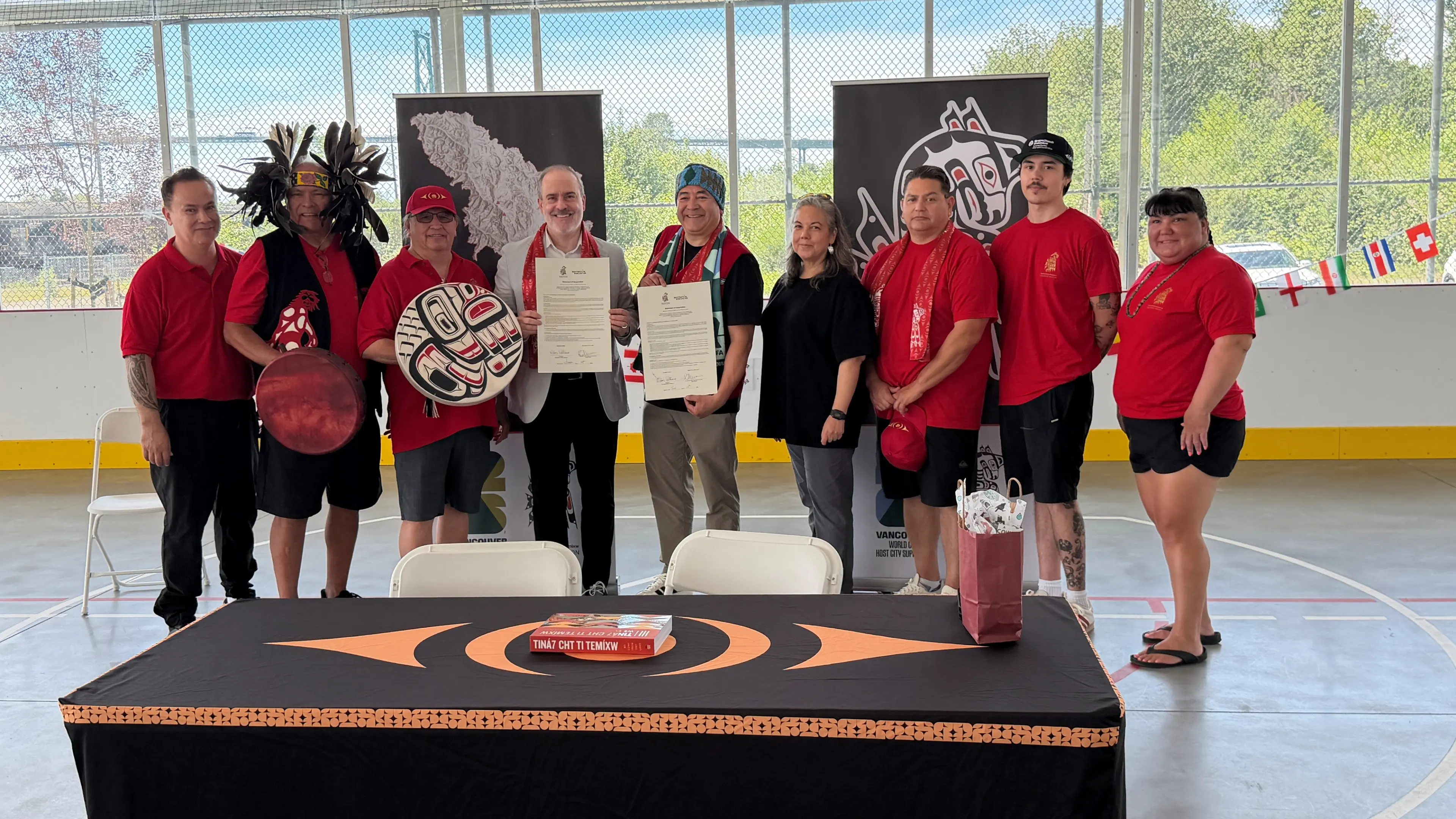 Group of ten people standing indoors behind a table with Indigenous art, two holding signed documents, some wearing red shirts and traditional regalia.