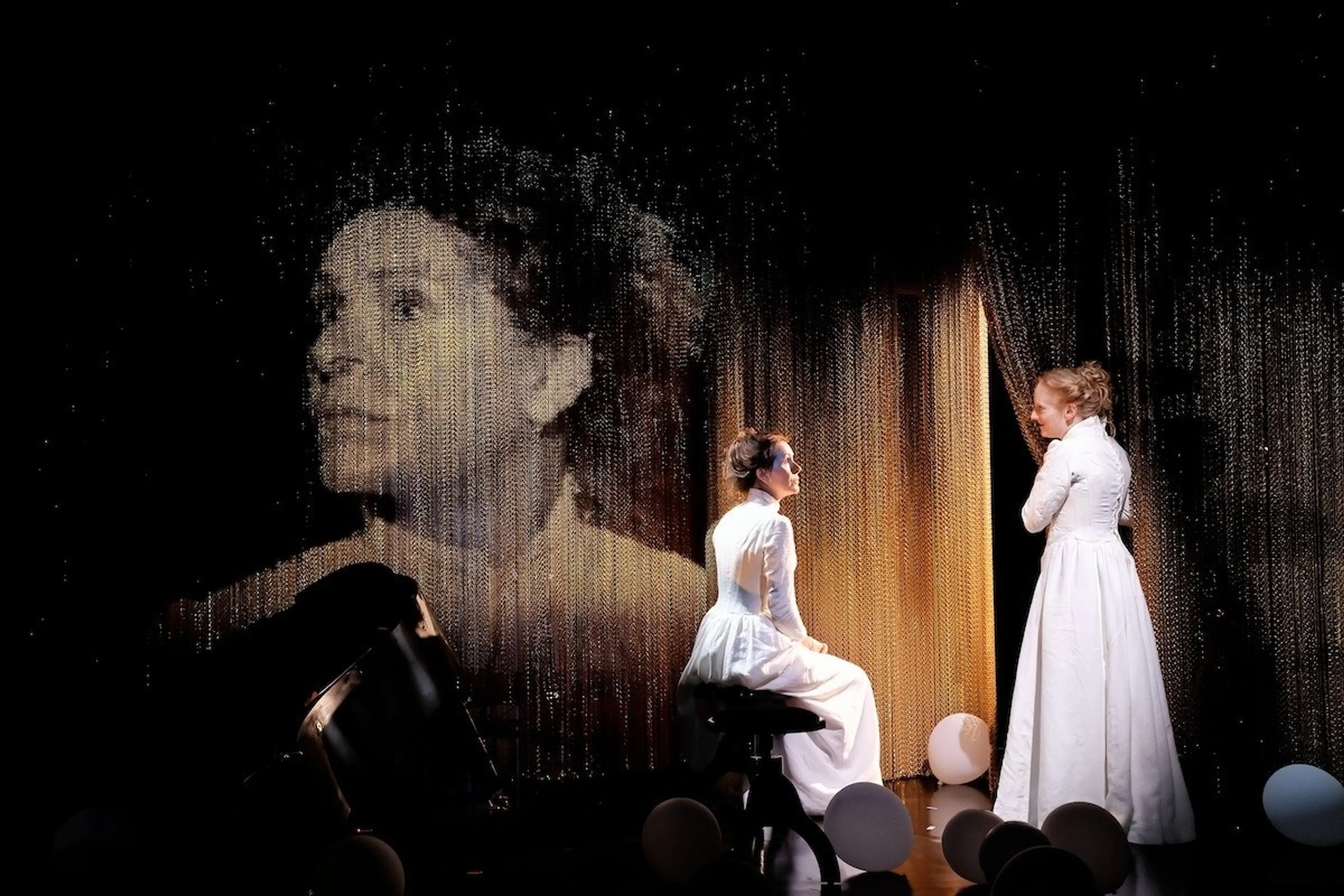 Two people in white period dresses on stage with a large projected face in the background and balloons on the floor.