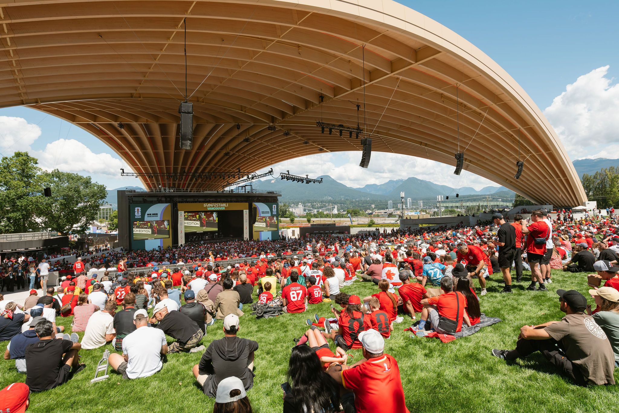 Large crowd wearing red sitting on grass under a curved wooden roof watching a stage with screens in an outdoor venue.