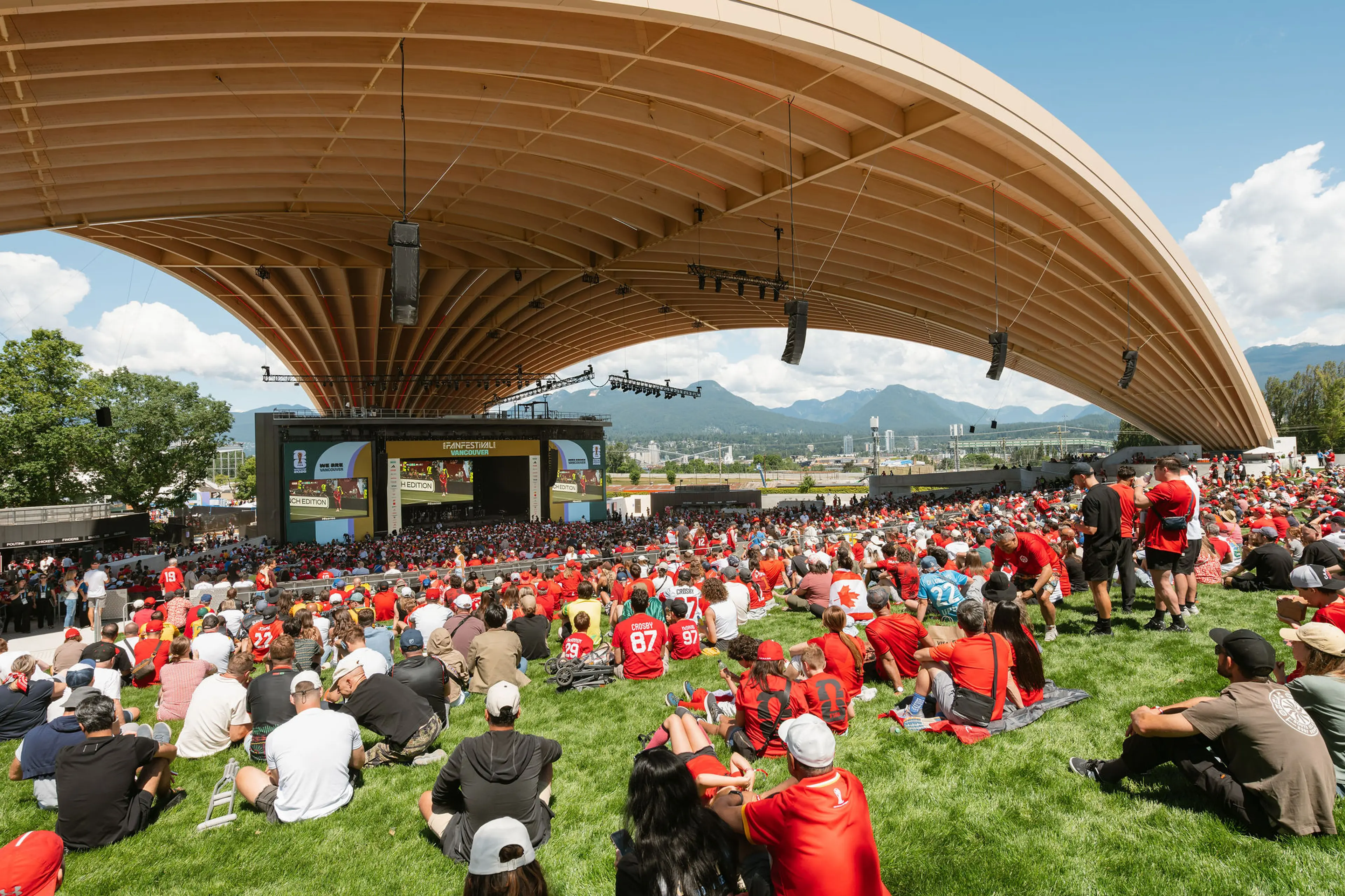 Large crowd wearing red sitting on grass under a curved wooden roof watching a stage with screens in an outdoor venue.