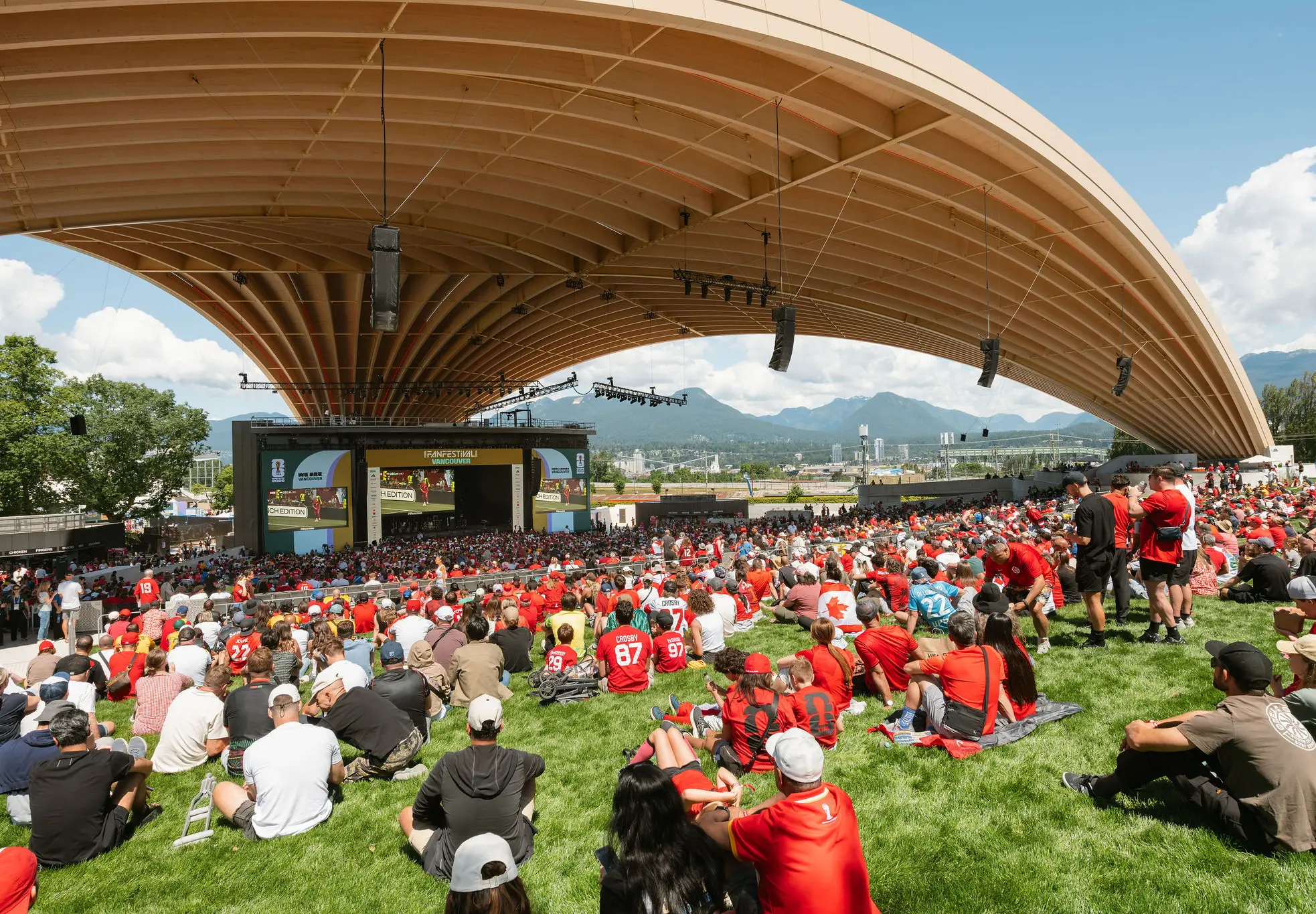 Large crowd wearing red sitting on grass under a curved wooden roof watching a stage with screens in an outdoor venue.