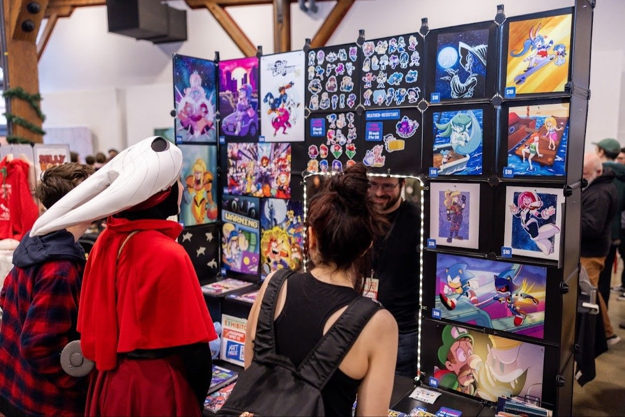 Three people looking at a display of colorful cartoon and video game art prints and stickers at a convention booth.