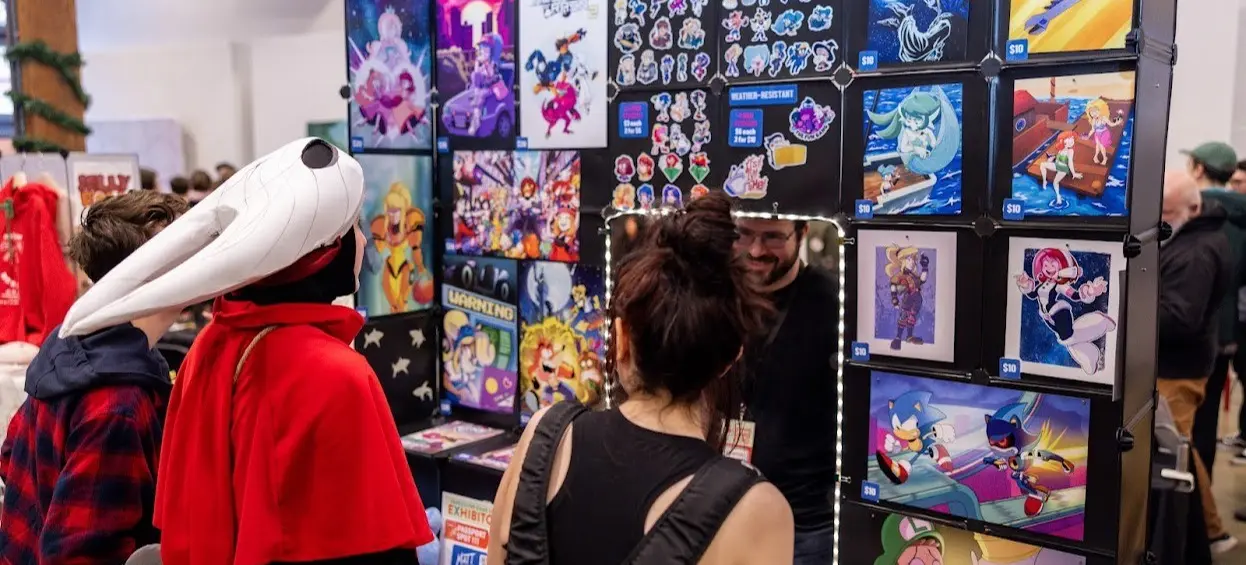 Three people looking at a display of colorful cartoon and video game art prints and stickers at a convention booth.