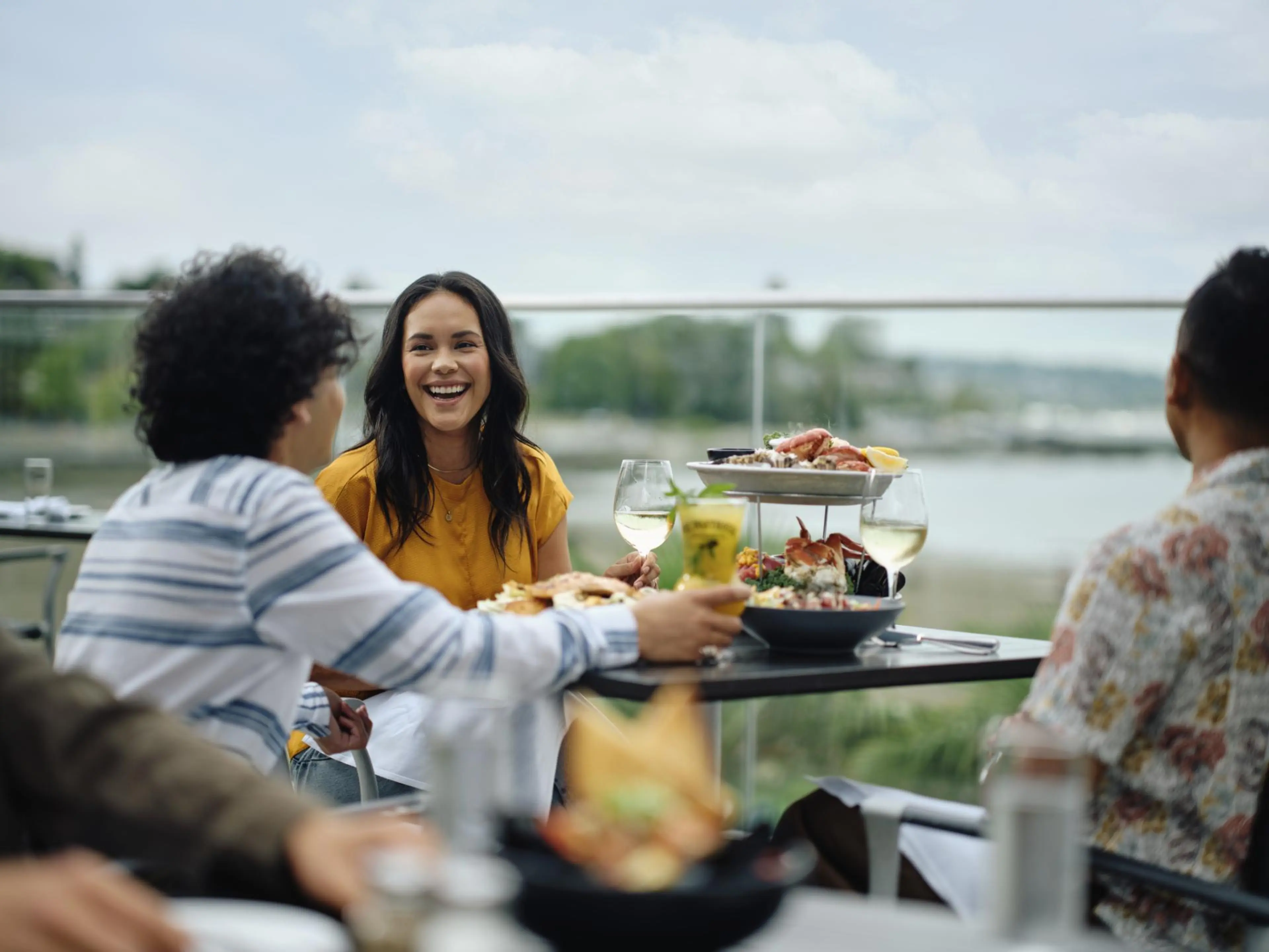 Three people sitting at an outdoor table with seafood dishes and drinks, one woman in a yellow top smiling.